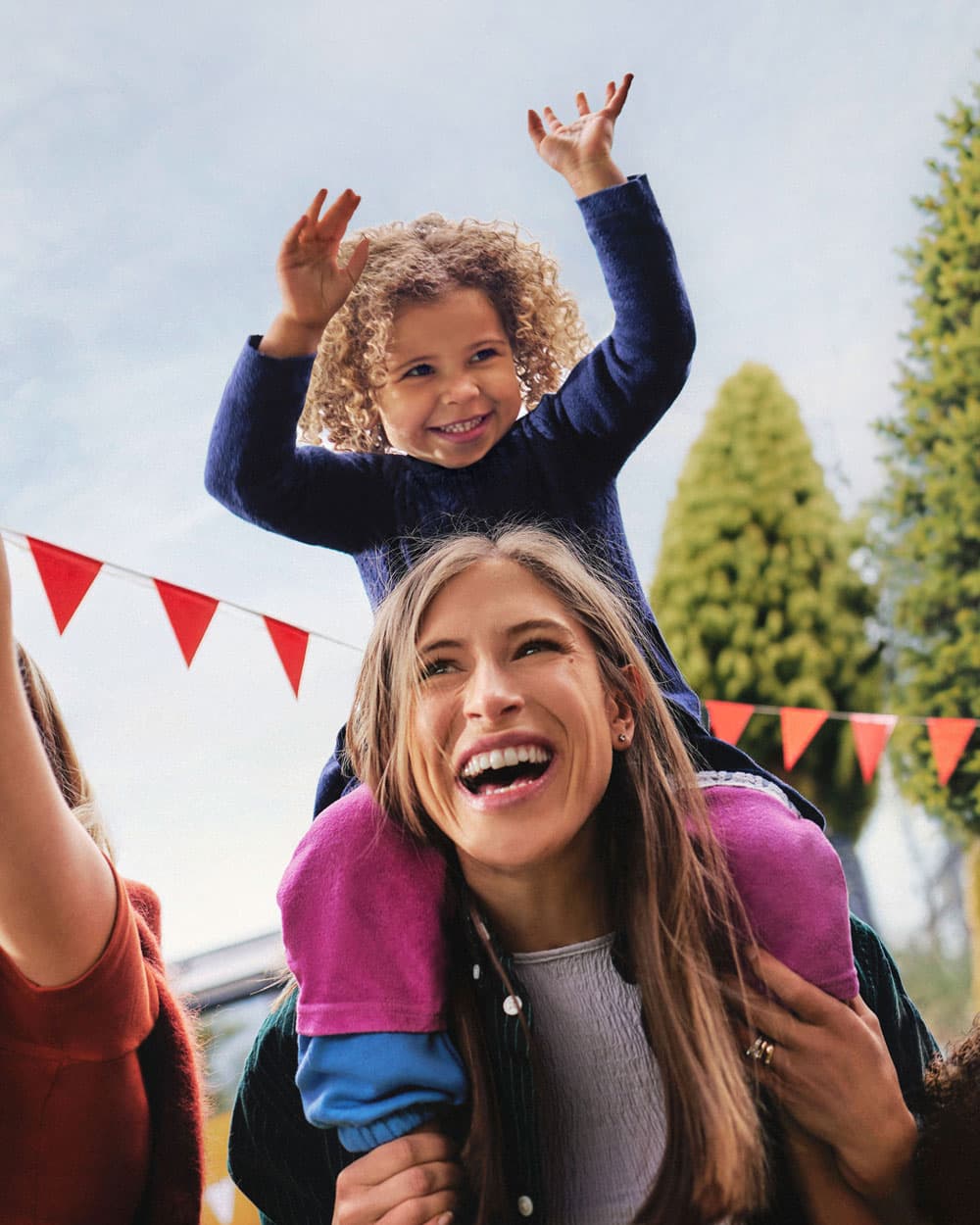Mutter tanzt auf einer Gartenparty mit ihrer Tochter auf den Schultern. Die fröhliche Szene steht für einen aktiven Lebensstil, Energie und Wohlbefinden im Familienalltag.