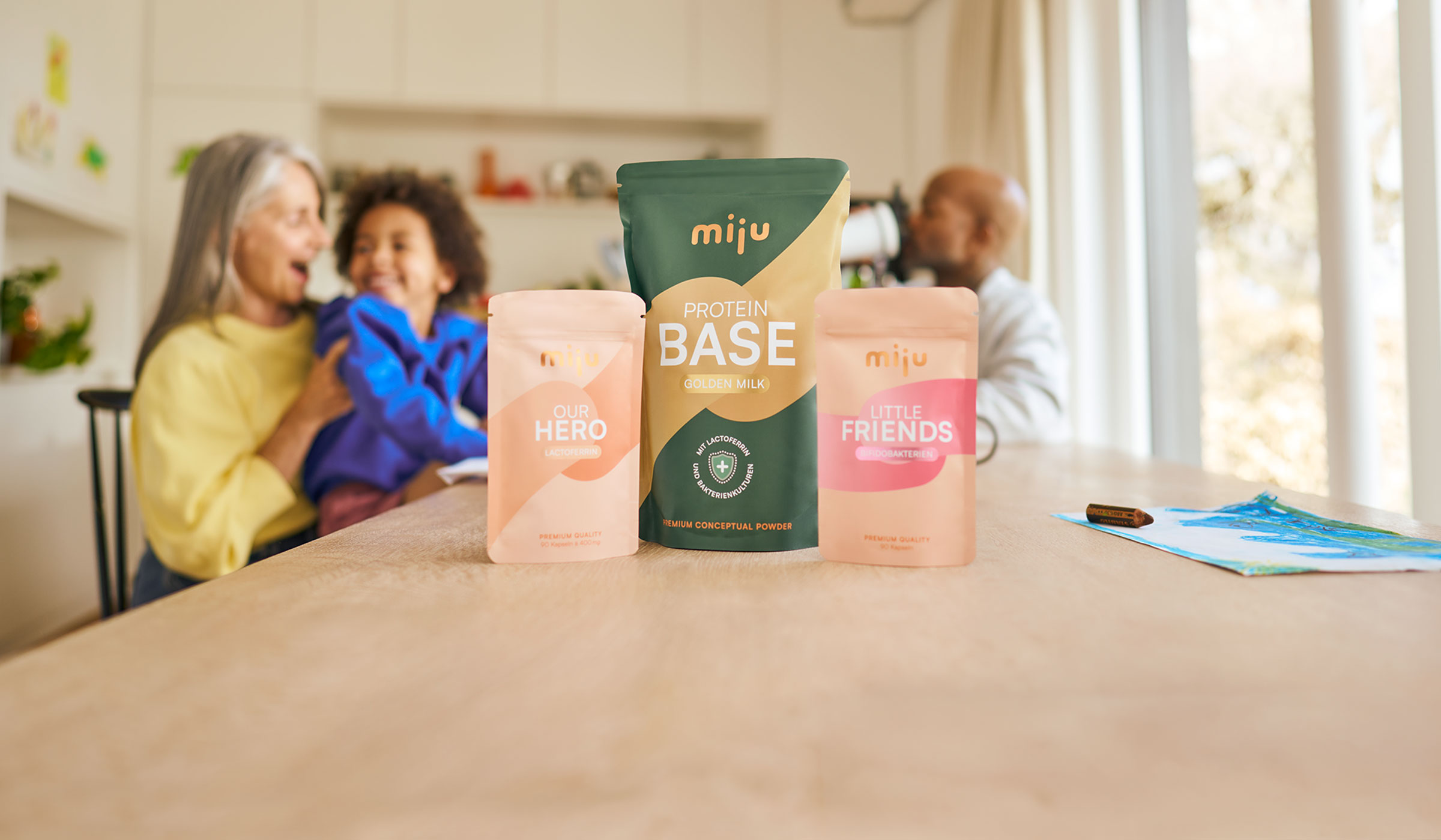 miju Lactoferrin, Bifidobakterien und Proteinpulver vor einer Familie im Alltag. Im Hintergrund trinkt ein Vater einen Proteinshake, während eine Großmutter gemeinsam mit ihrem Enkel lacht.