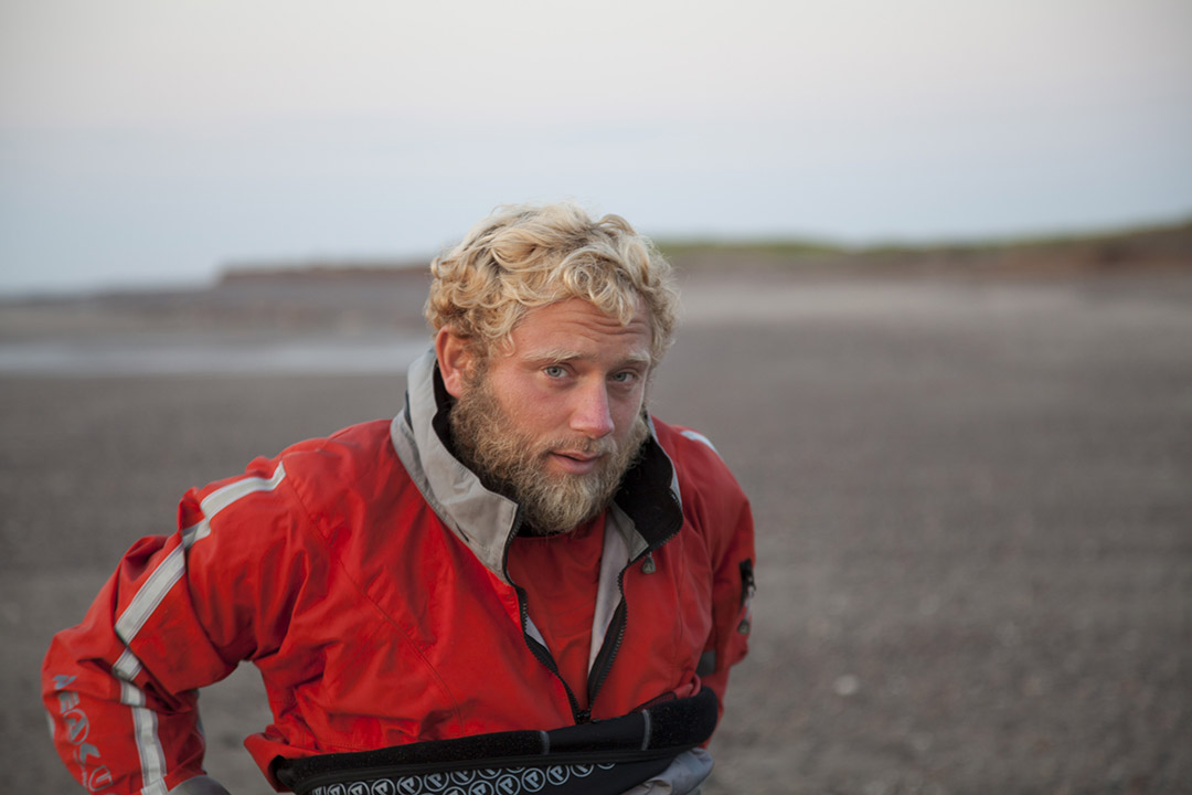Bearded man with curly blond hair wearing a red waterproof jacket on a rocky beach.