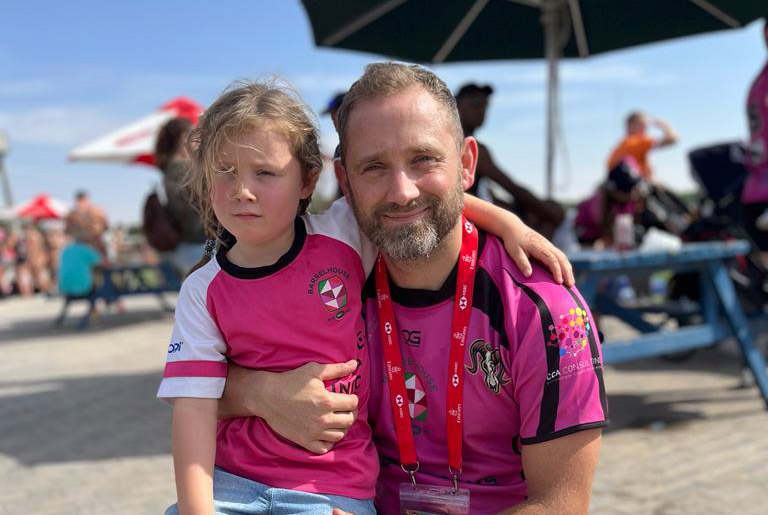 Man with beard smiling and hugging a young child wearing matching pink sports jerseys outdoors.