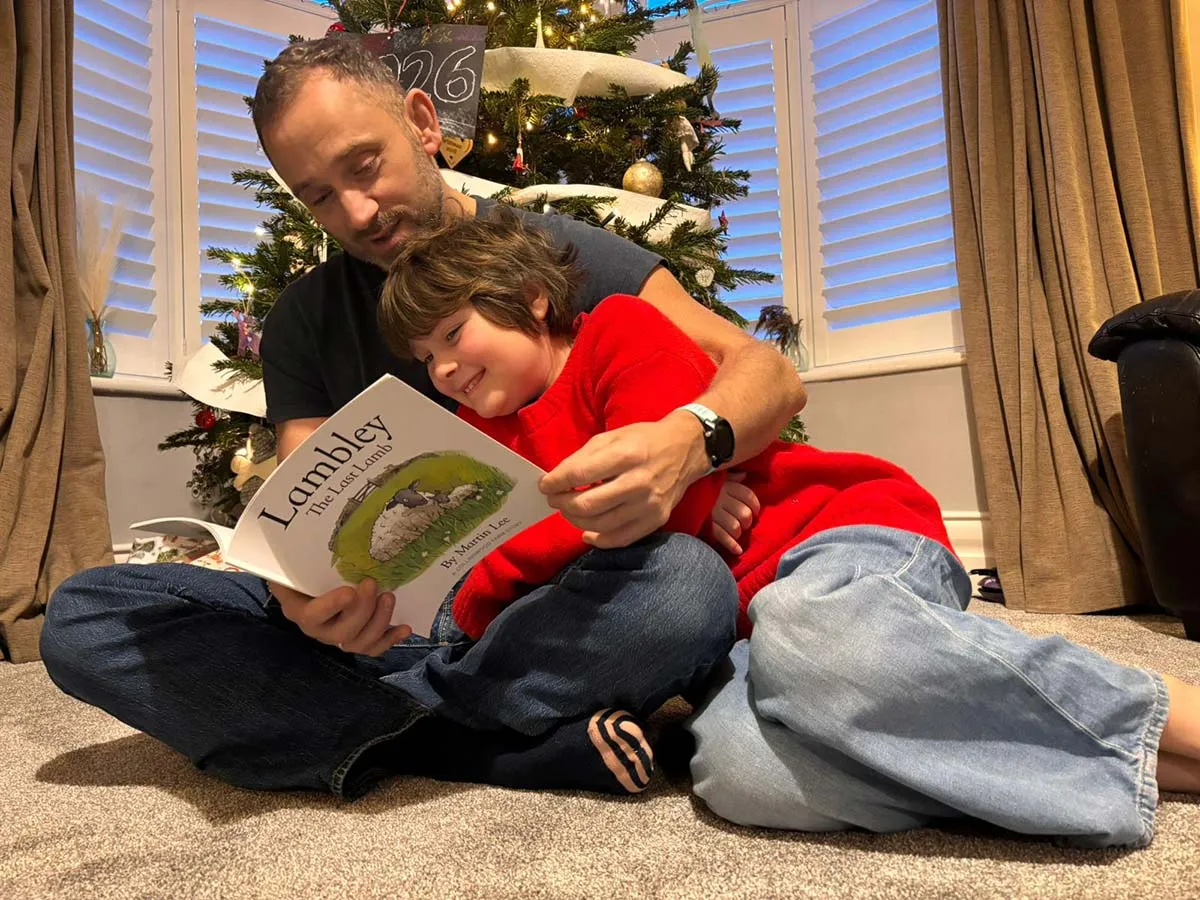 Man and child sitting on carpeted floor in front of a decorated Christmas tree, reading the book 'Lambley The Lost Lamb' together.