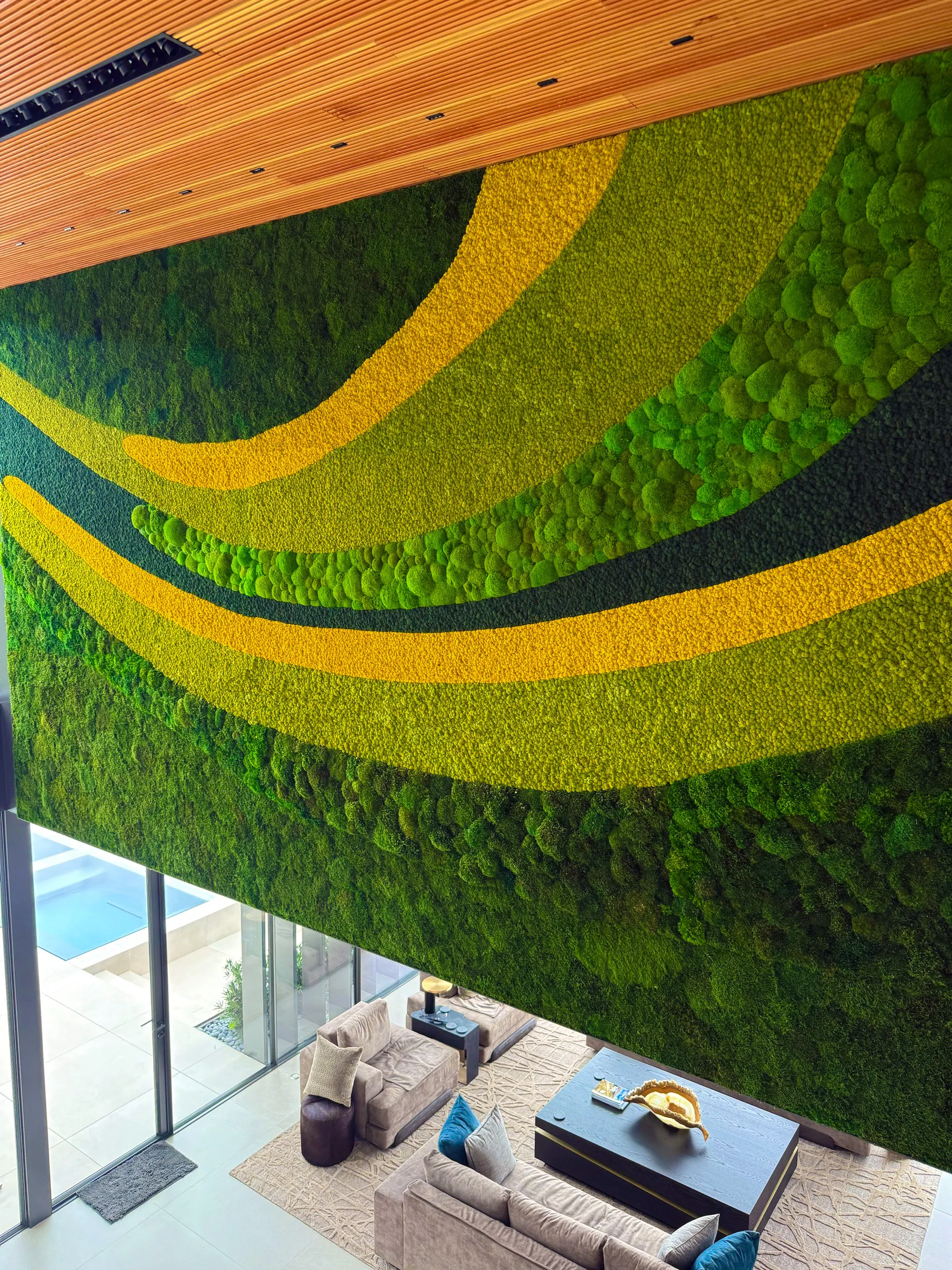 Two-story preserved moss feature wall with flowing gold, lime and pillow moss bands above a living room with a pool view, by Austin Moss Creations