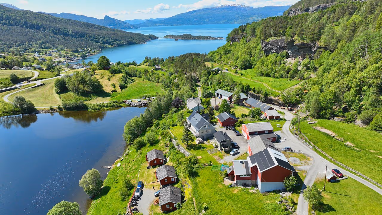 Liten norsk landsby med røde hus nær en innsjø og fjell med frodig grønt landskap under en blå himmel.