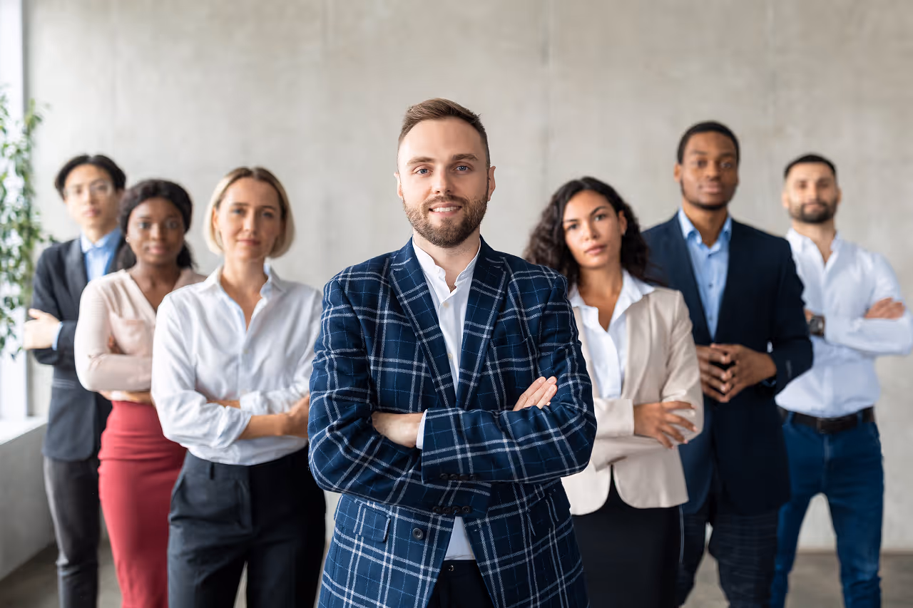 Egy fiatal férfi keresztbe tett karokkal áll középen, mögötte hat különböző nemzetiségű üzleti öltözetű ember áll sorban.