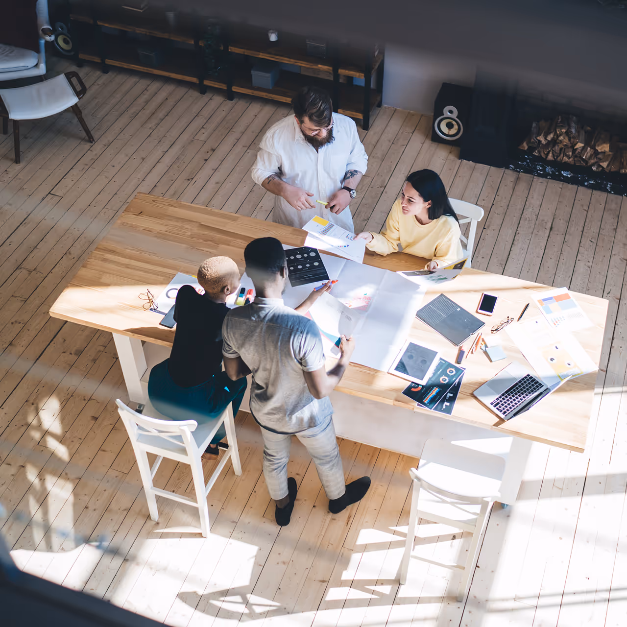 Négy ember egy faasztal körül egy világos szobában, megbeszélésen vagy projekten dolgoznak papírokkal és laptopokkal.
