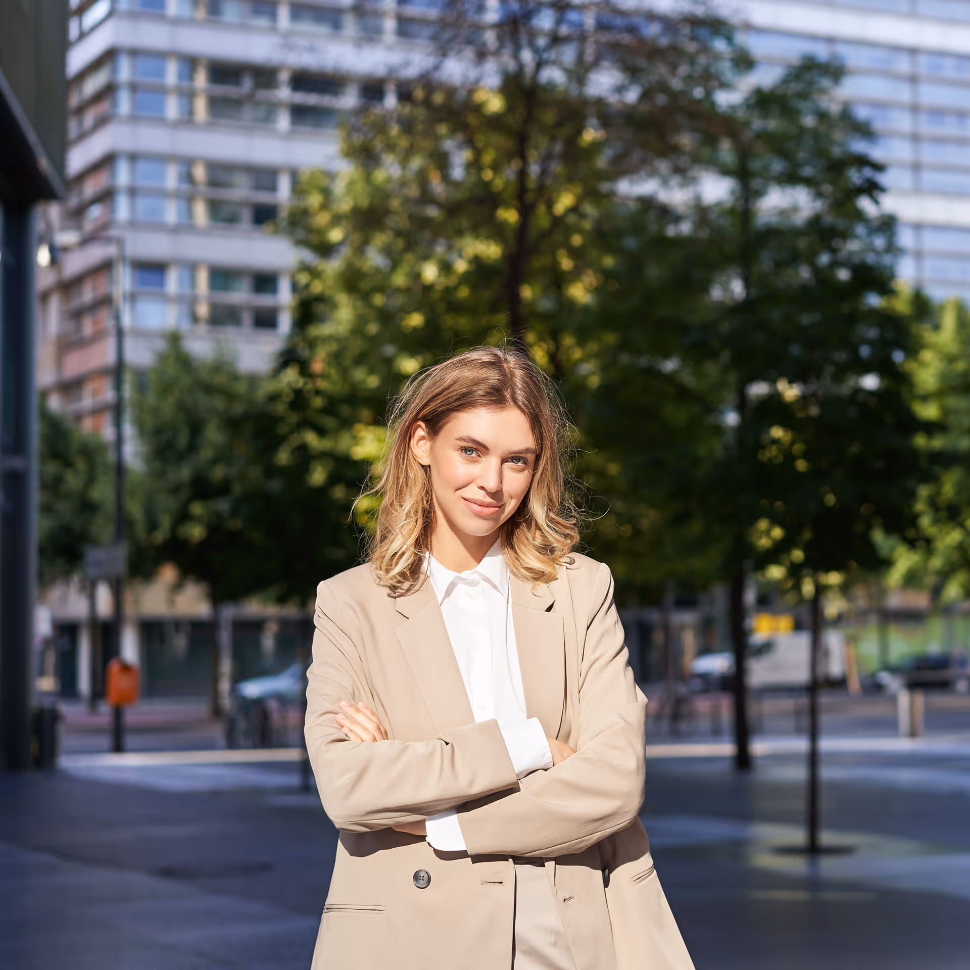 Mosolygó fiatal nő keresztbe font karokkal, bézs öltönyben városi környezetben.