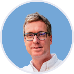 Mosolygó férfi szemüvegben fehér ingben világoskék háttérrel.