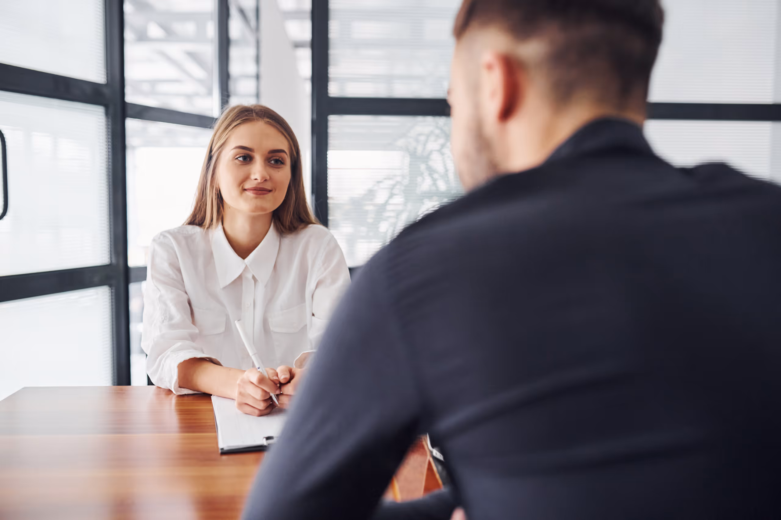 Egy fiatal nő fehér ingben jegyzetel egy interjún egy férfival, aki háttal ül egy modern irodában.