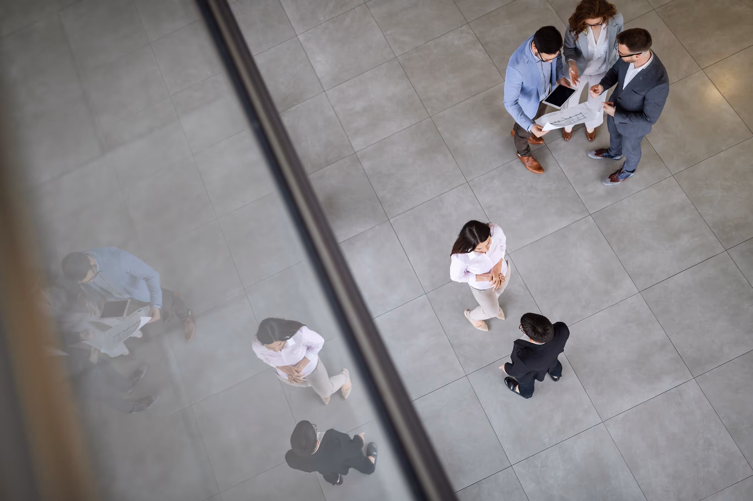 Üzletemberek beszélgetnek és néznek terveket egy nagy mozaikpadlós térben felülnézetből, egyikük tabletet tart.