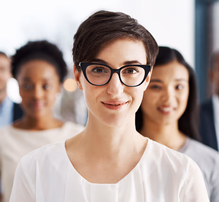 Középen álló rövid hajú nő fekete szemüveggel, mögötte mosolygó emberek homályosan.