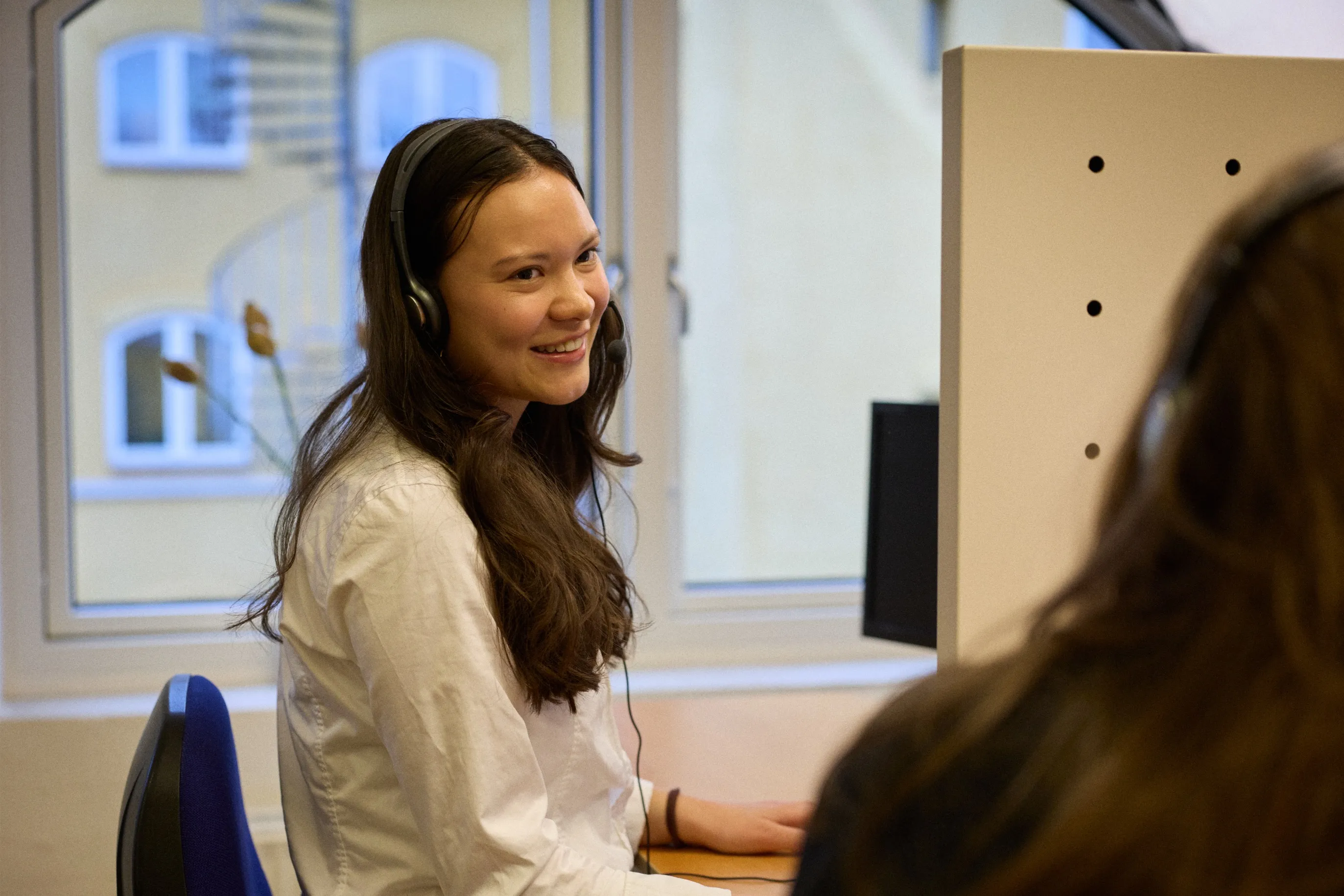 Smiling woman with long hair wearing a headset and white shirt sitting at a desk in front of a window.