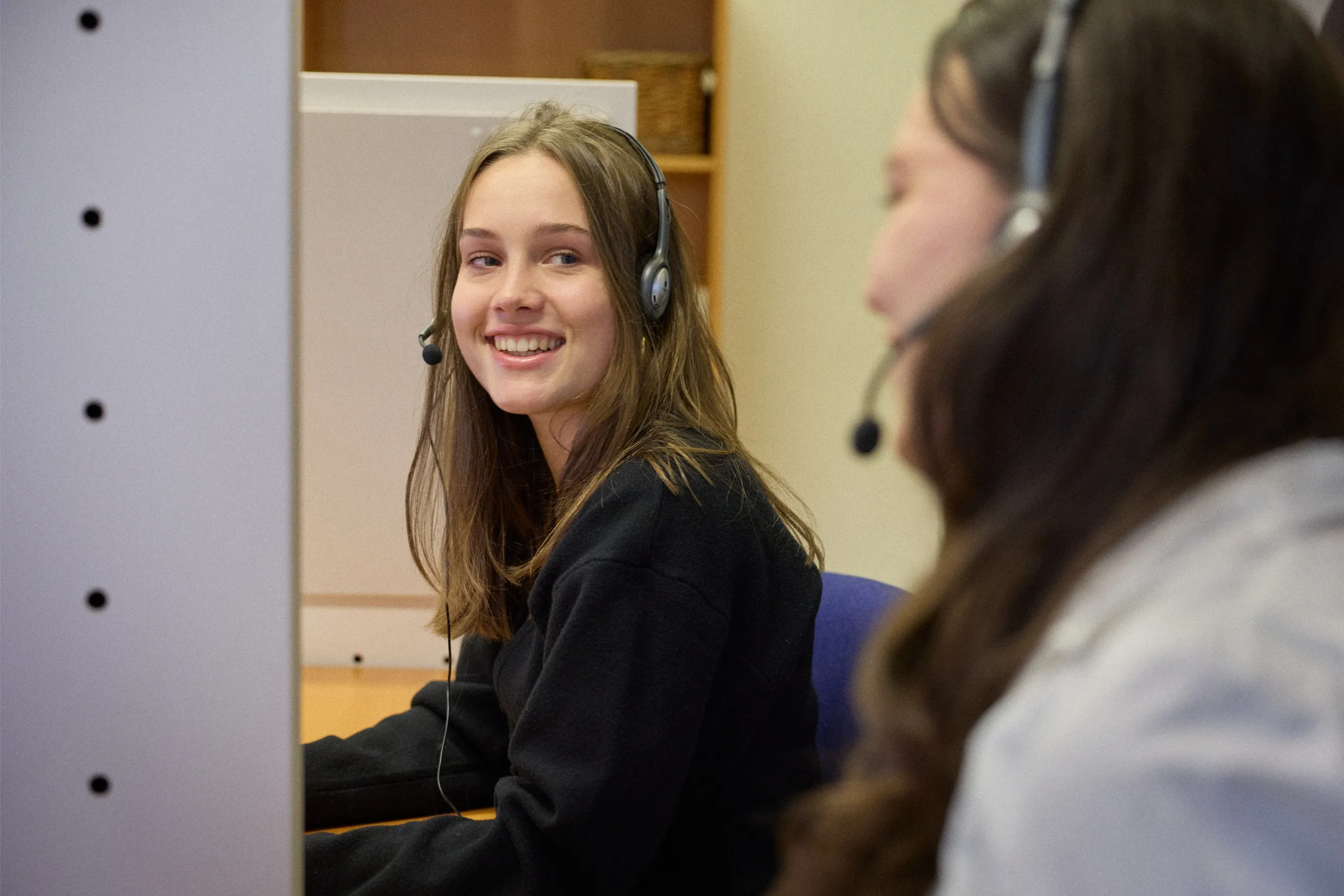 Two women wearing headsets working together in an office, one smiling and looking at the other.