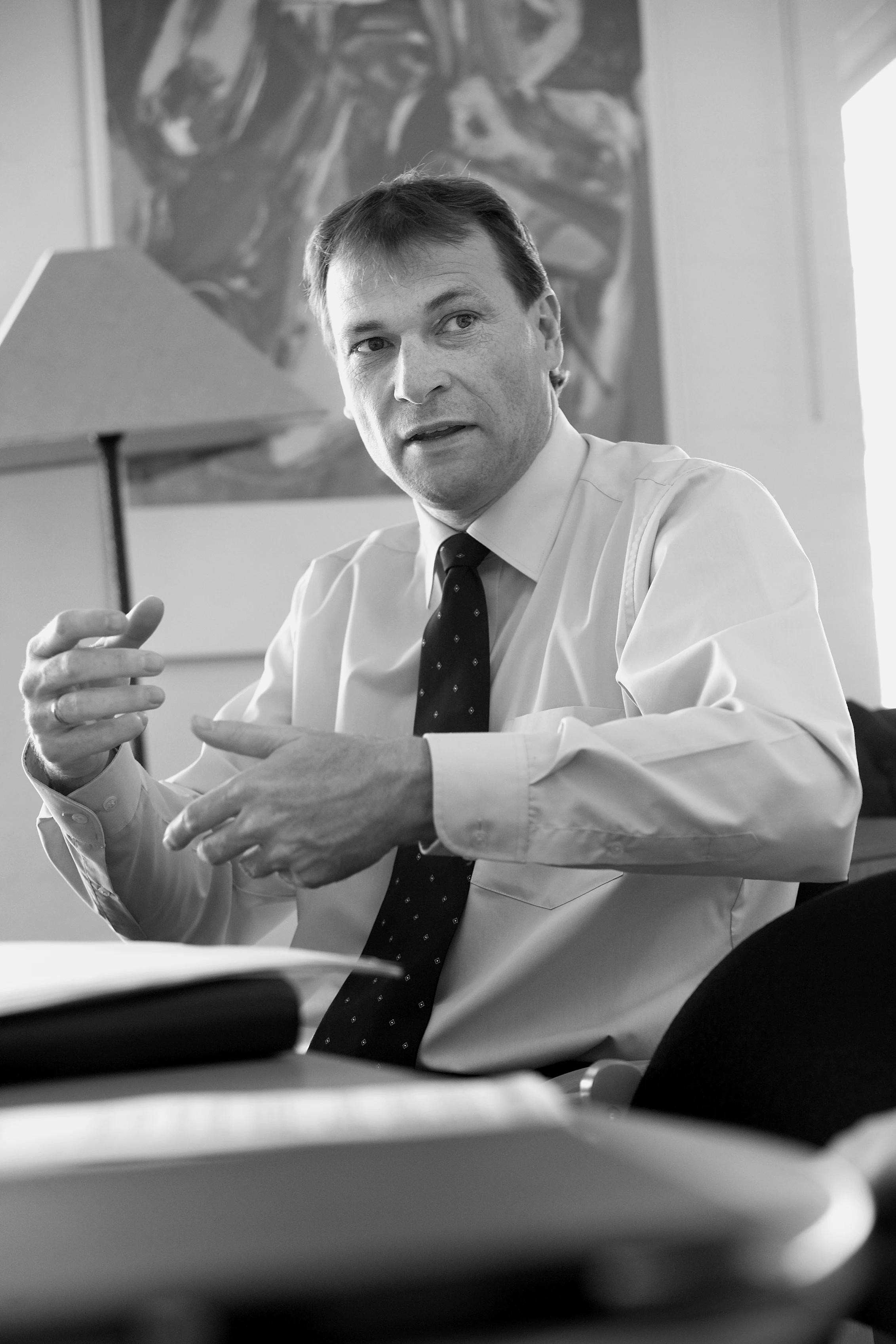 Man in a blue striped shirt sitting at a desk holding a red Ferrari toy car with office decor and a laptop nearby.