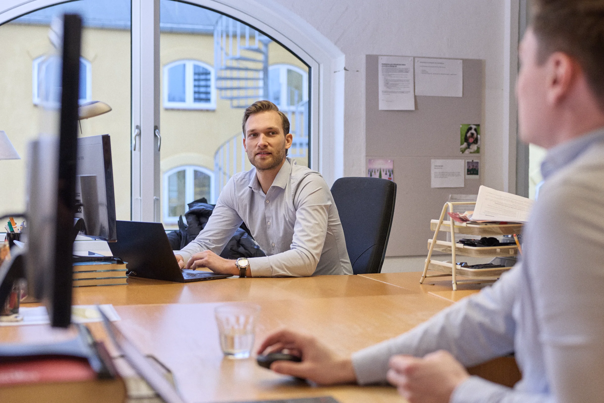 Three men in a meeting room with laptops and notebooks, two sitting and one standing near a whiteboard.