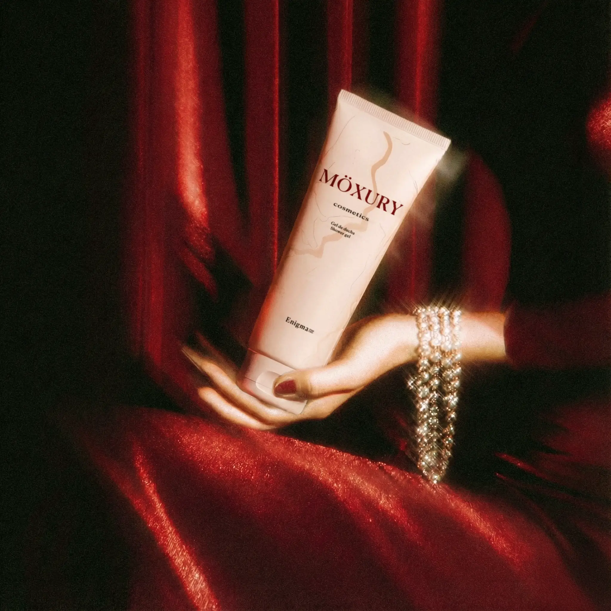 Gel de ducha Moxury Cosmetics sostenido por una mano femenina sobre fondo de tela roja satinada, imagen de cosmética de lujo.