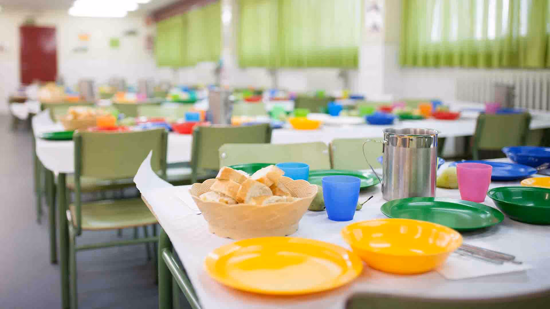 Empty school cafeteria with neatly set tables, colorful plates and cups, and a basket of bread on display.