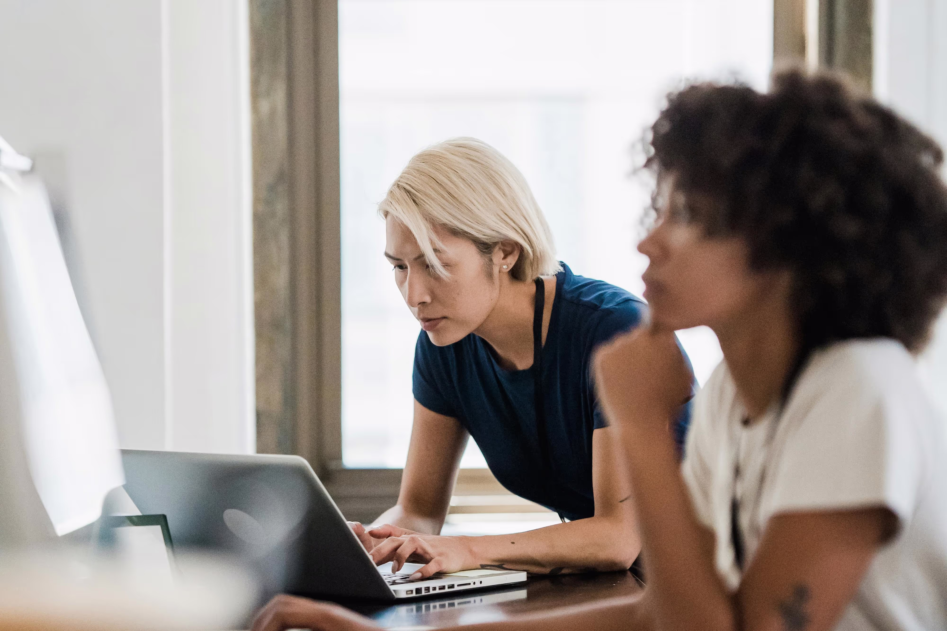 Two women at a desk, one with blonde hair focused on typing on a laptop, the other with curly hair resting her chin on her hand.