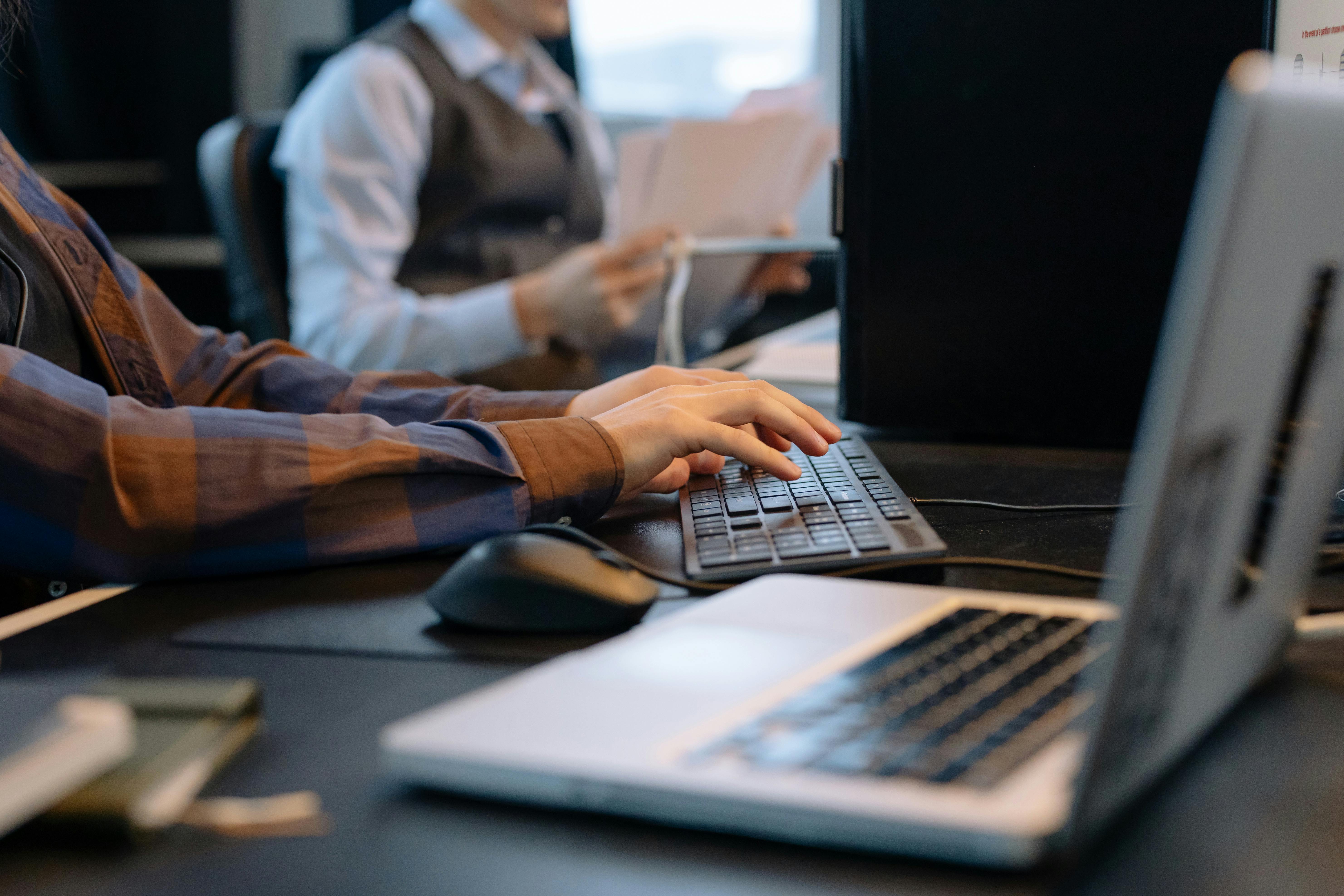 Person typing on a keyboard at a desk with a laptop in the foreground and another person holding papers in the background.
