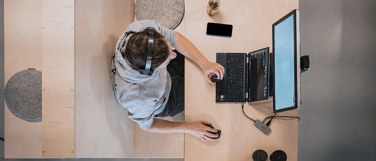 En person med headset ved et skrivebord, set ovenfra, arbejder på en bærbar og en ekstra skærm.