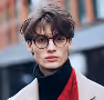Young person with short brown hair, wearing round glasses, a black turtleneck, and a red and beige jacket, standing outdoors.