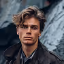 Portrait of a young man with tousled hair wearing a dark jacket, standing outdoors near rocky terrain.