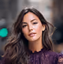 Portrait of a woman with long dark hair wearing a purple lace top, posing outdoors with a blurred urban background.