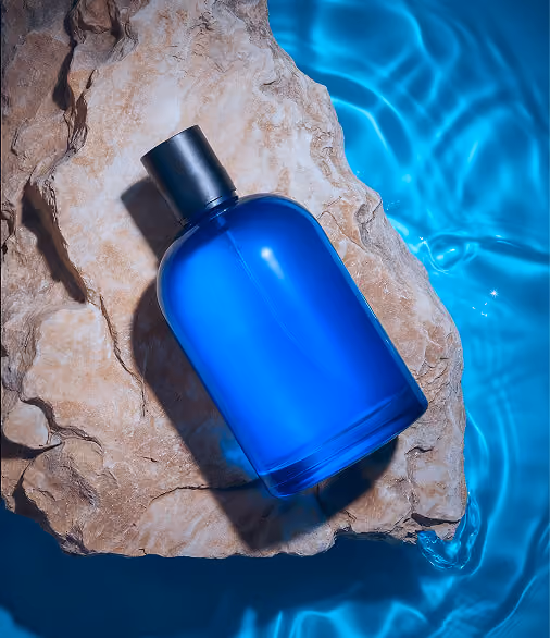 Blue perfume bottle with black cap resting on textured rock beside rippling blue water.