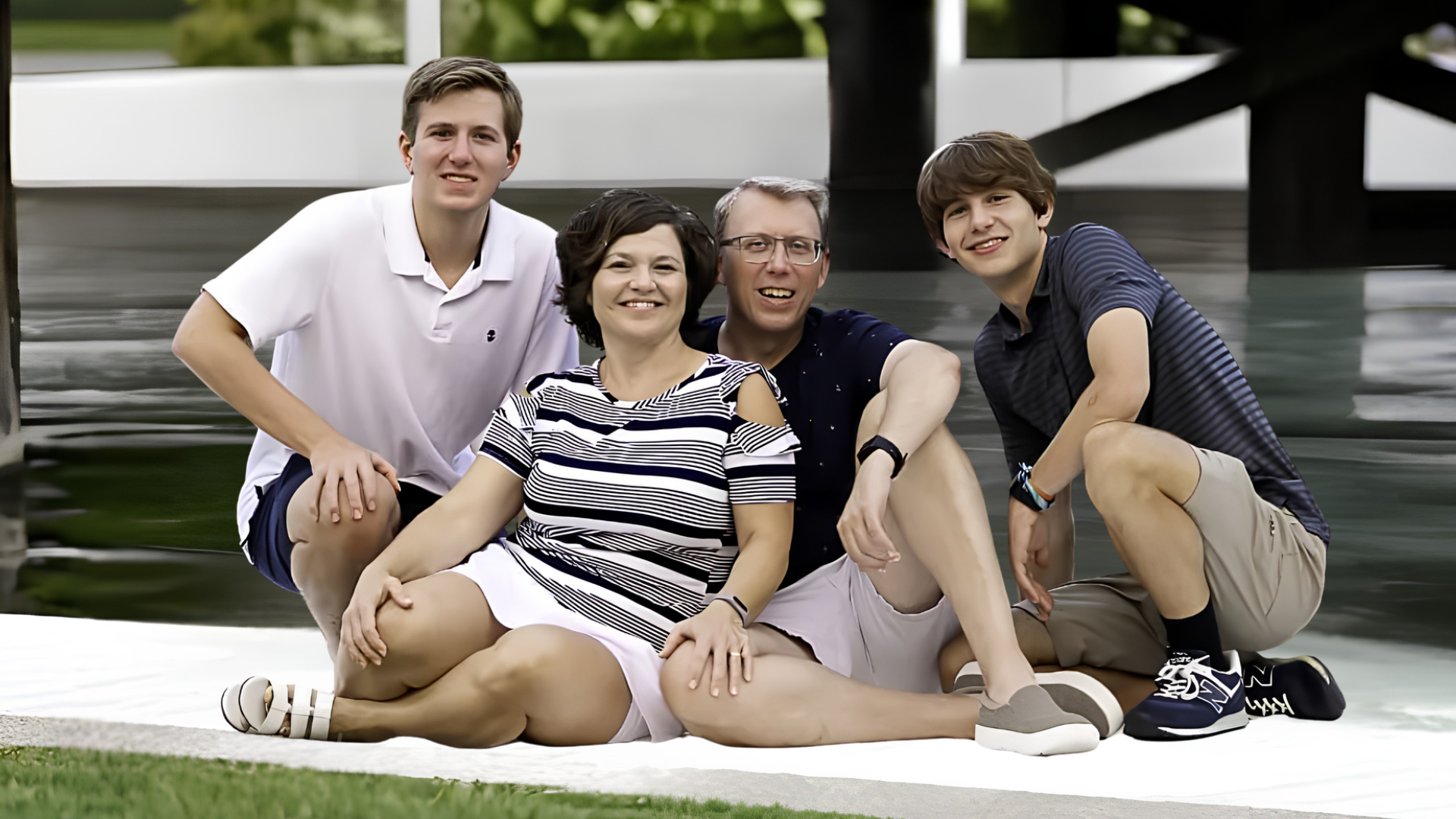 Family of four smiling together outdoors near a waterfront, enjoying quality time in a relaxed setting with modern architecture and greenery in the background.