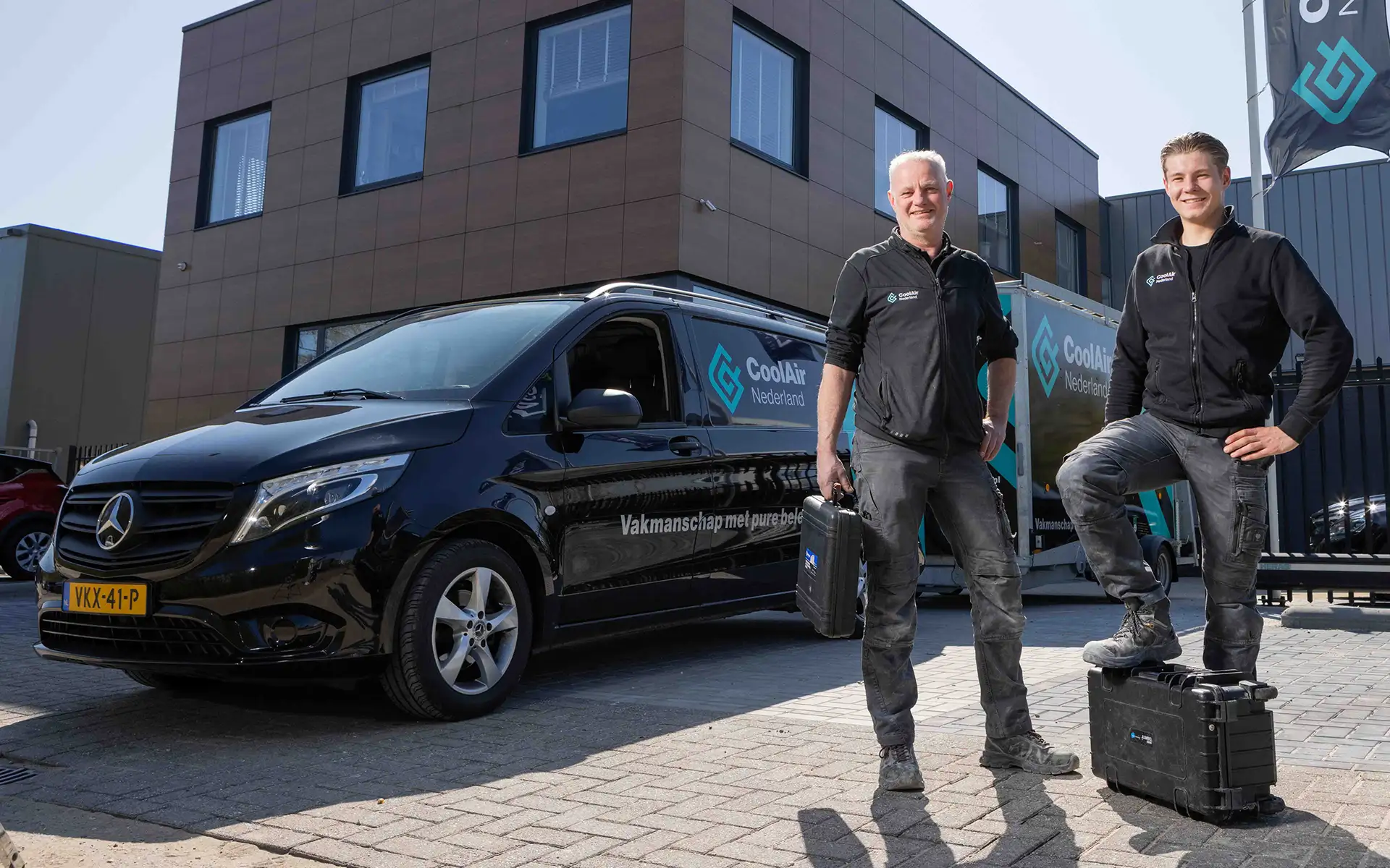 Twee mannen in werkkleding poseren naast een zwarte bedrijfsbus en een aanhanger met het logo van CoolAir Nederland.