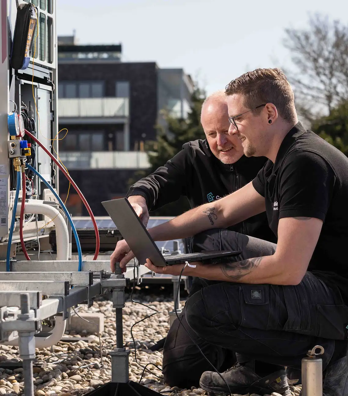 Twee technici controleren buiten een HVAC-installatie met een laptop op een zonnige dag.