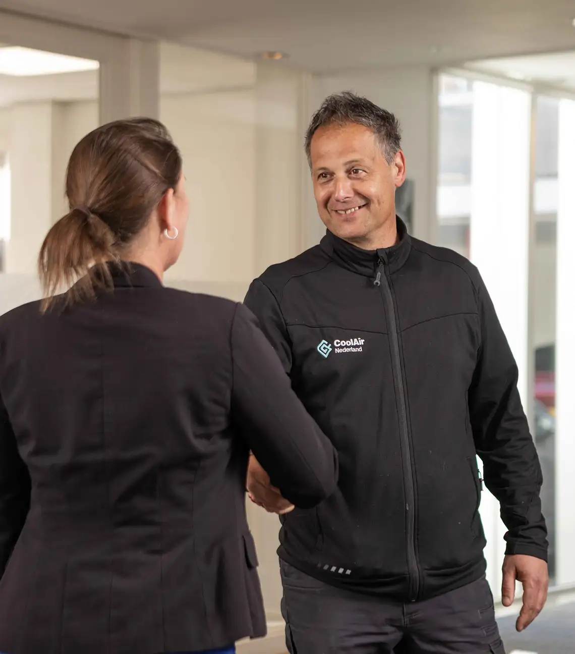 Een man met een zwart jasje met het logo 'CoolAir Nederland' geeft een hand aan een vrouw in een zwart colbert.