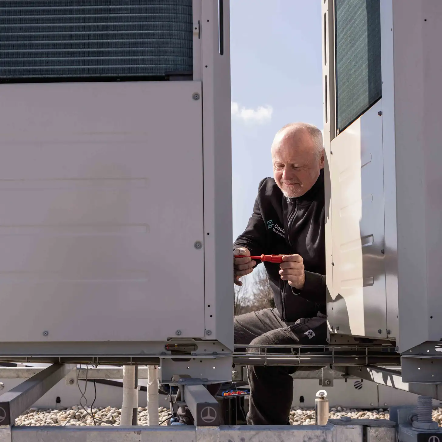 Technicus aan het werk aan een airconditioningsysteem op een dak onder een blauwe lucht.