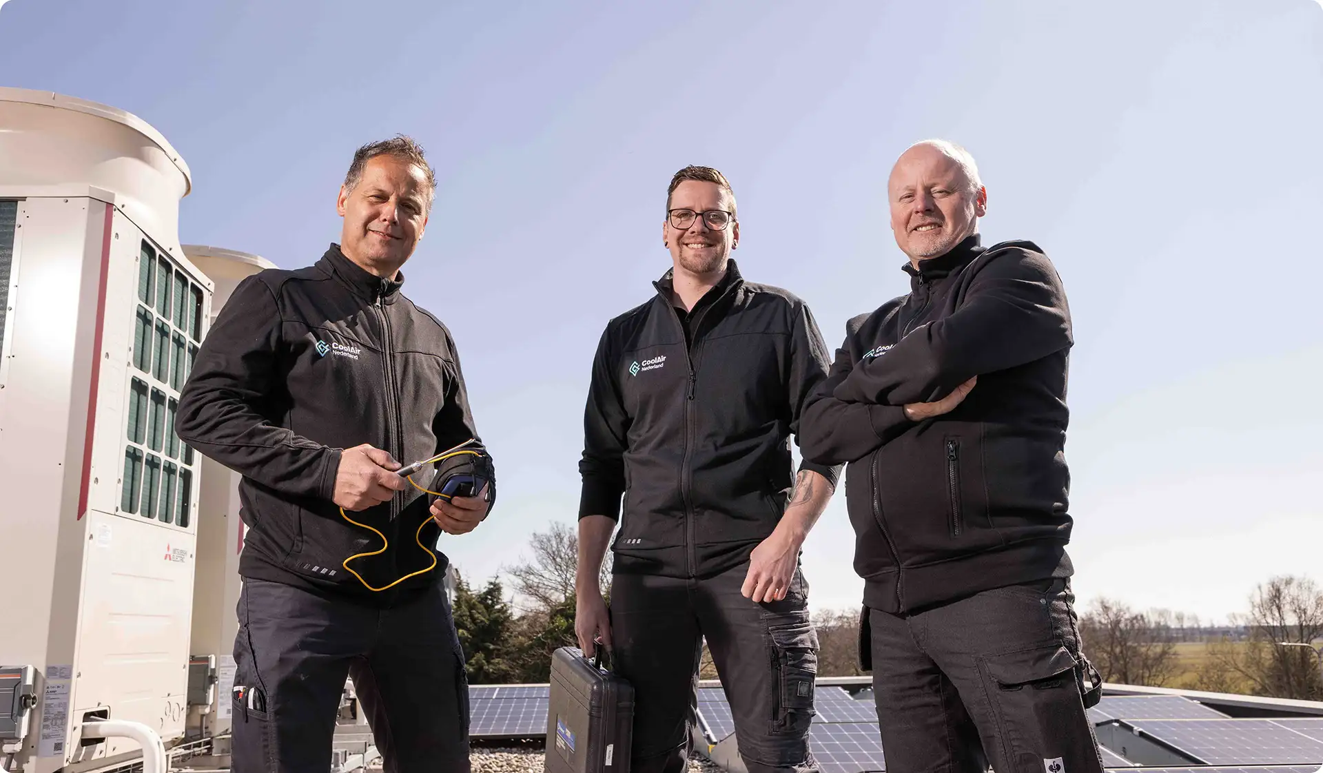Drie mannen in zwarte werkkleding met CoolAir Nederland-logo poseren buiten bij airconditioningunits en zonnepanelen.