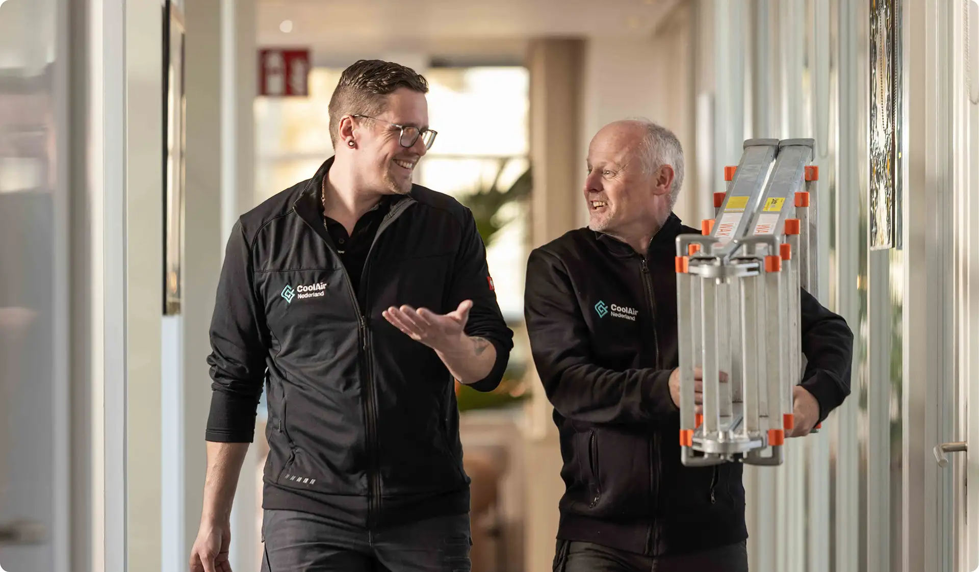 Twee mannen in zwarte jassen met het CoolAir Nederland-logo lopen in een gang, waarbij de oudere man een opgevouwen ladder draagt.
