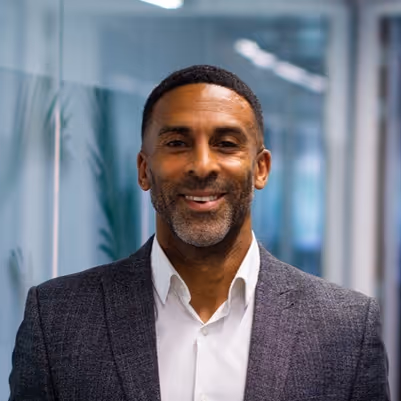 Portrait of Sean Barker wearing a grey blazer and white shirt, standing indoors with glass walls in the background.