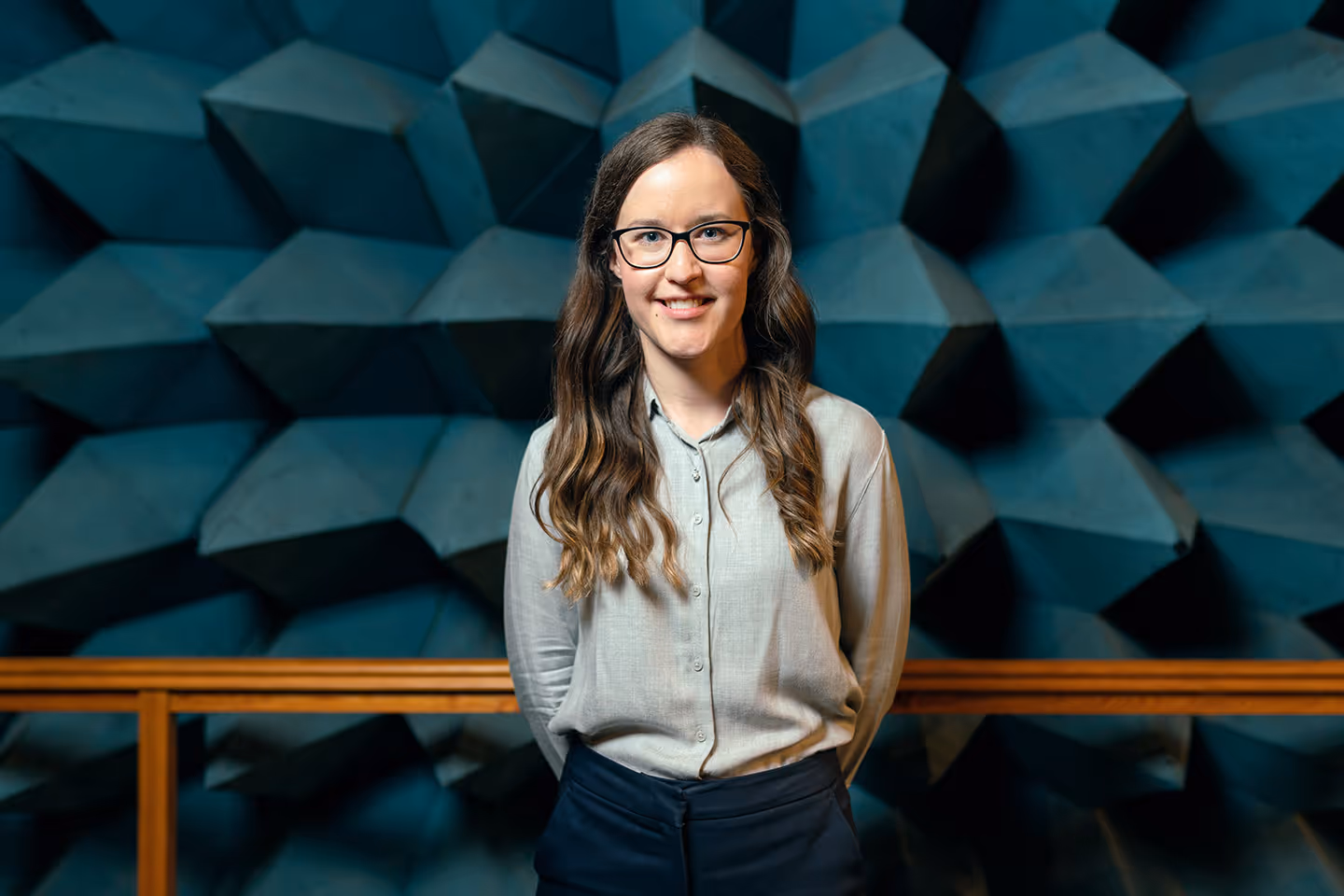 Woman with glasses and long wavy hair smiling, standing in front of a blue geometric patterned wall.
