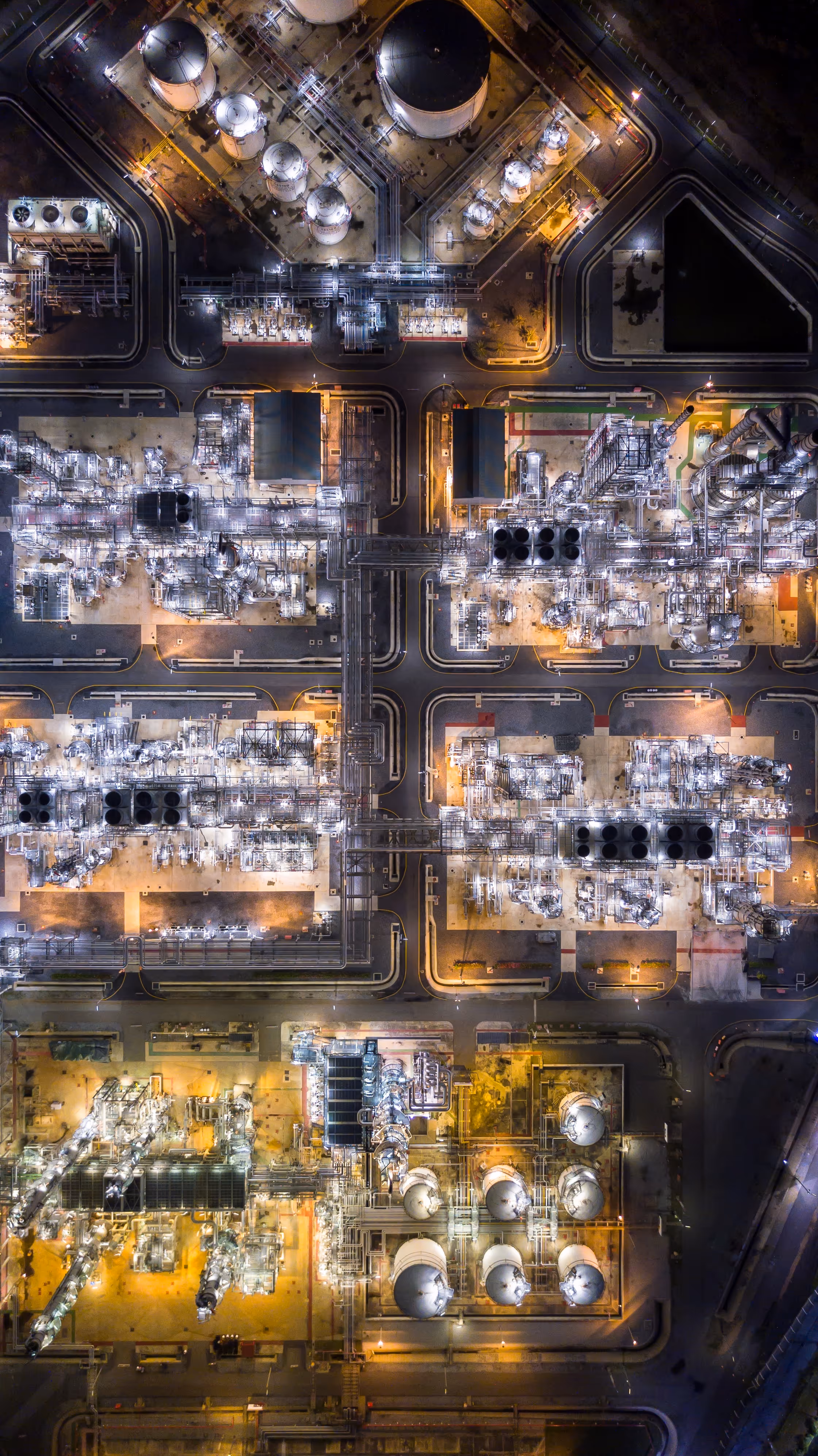 Aerial night view of an illuminated industrial refinery with storage tanks, pipelines, and complex machinery.