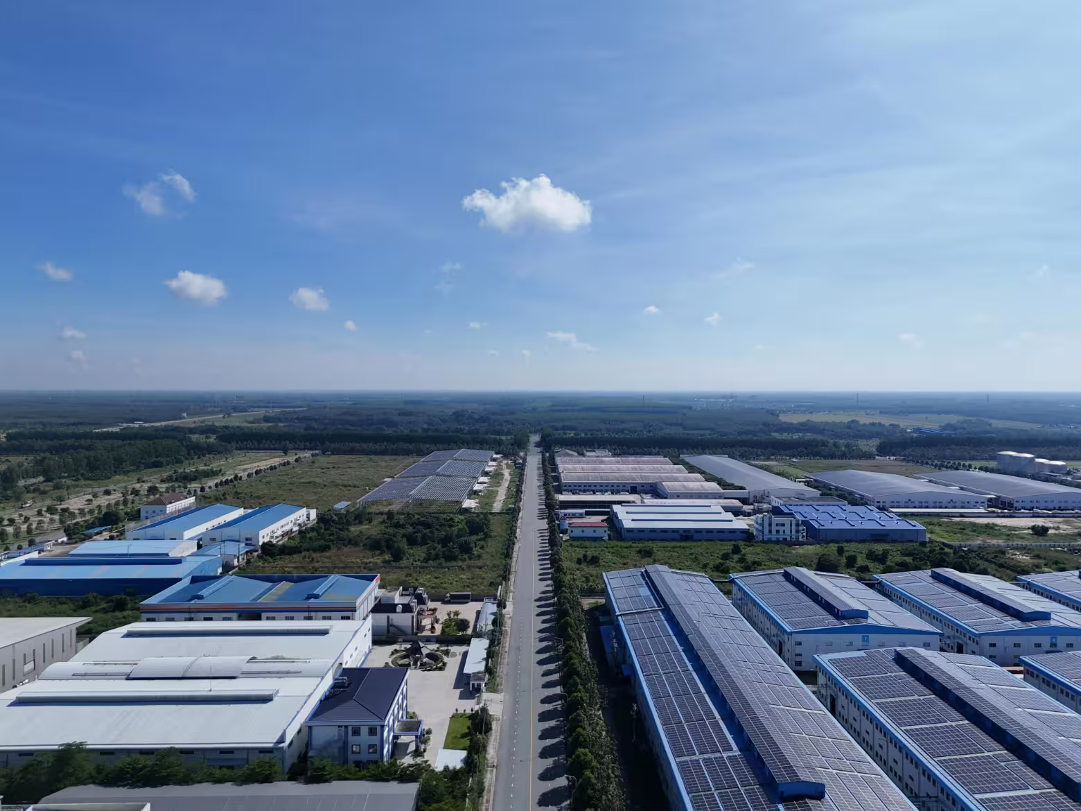 Aerial view of an industrial area with large factories, solar panels on roofs, and a long road lined with trees leading into the distance under a blue sky with scattered clouds.