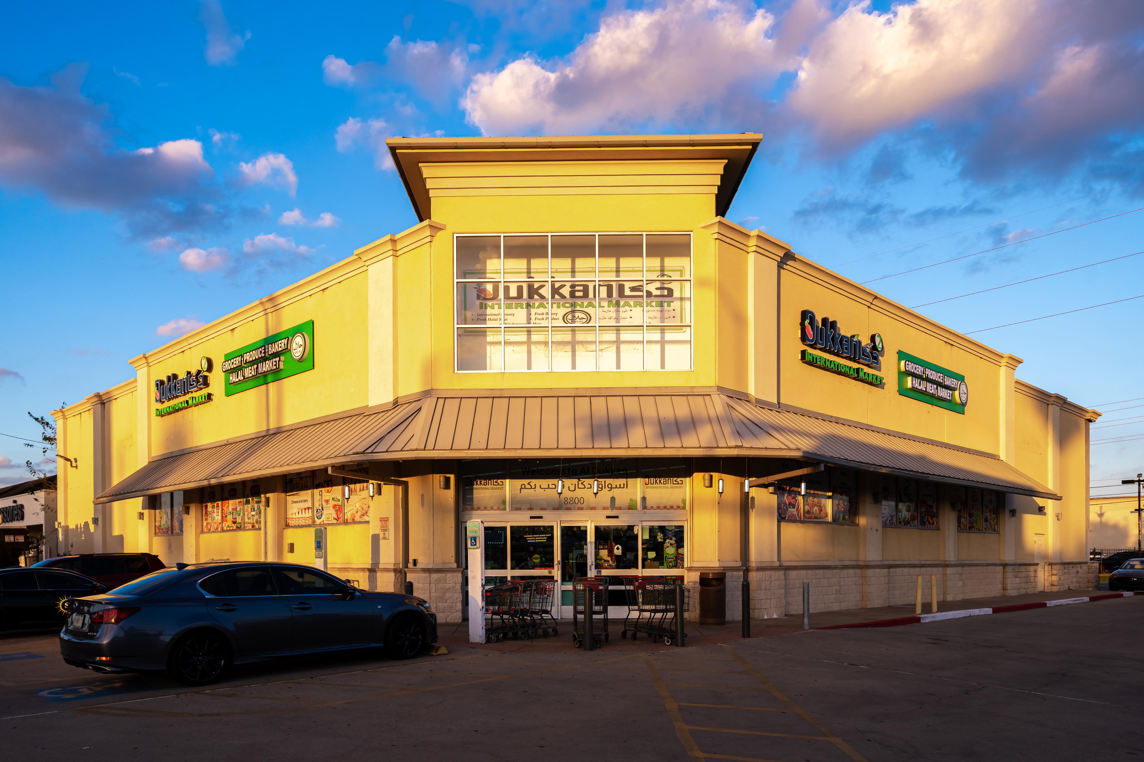 Aerial view of a parking lot in front of a grocery store named Sukkari's with several cars parked and city skyline in the distance under a blue sky.