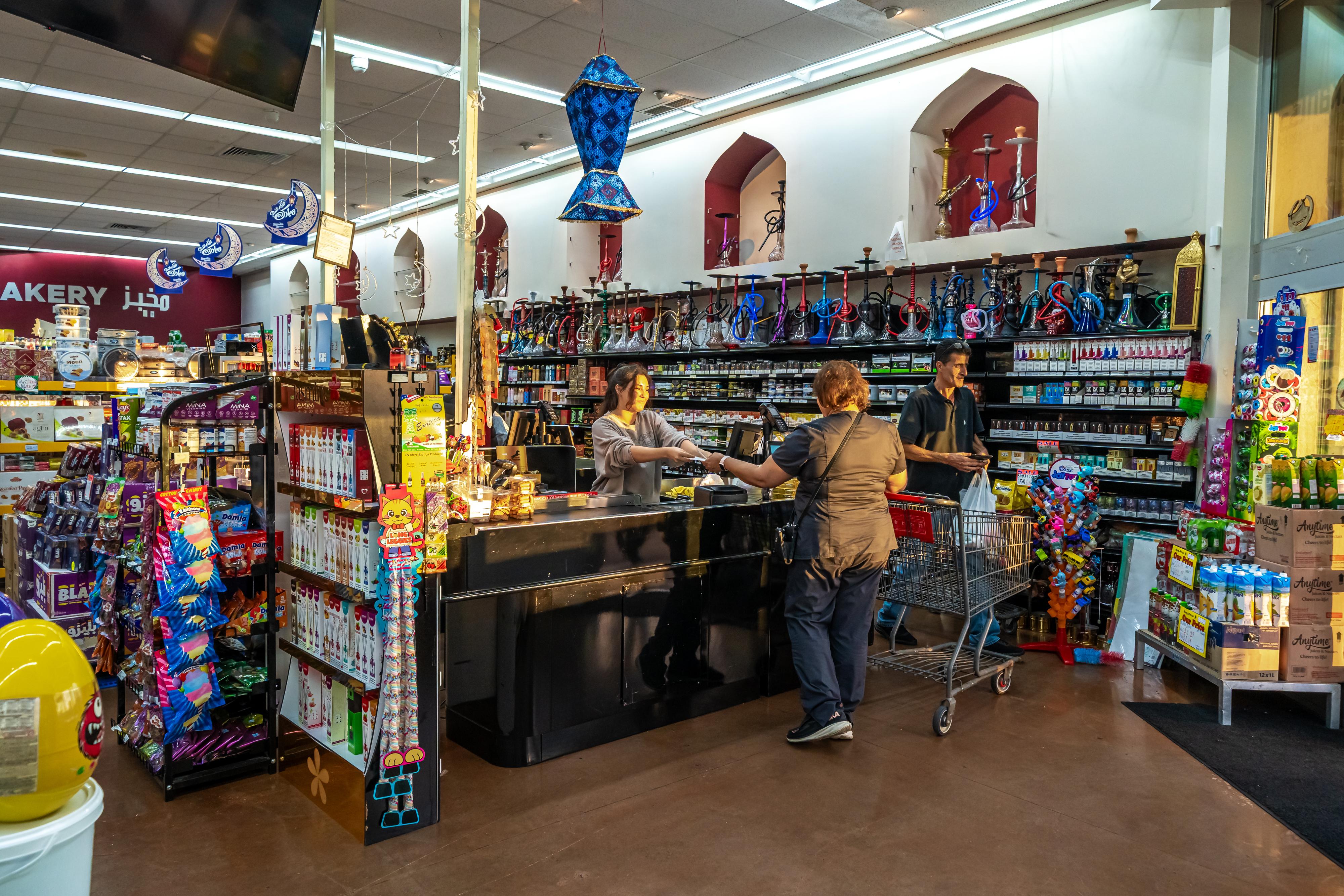 Customer paying at checkout counter in a store selling hookahs and groceries, with a man pushing a shopping cart nearby.