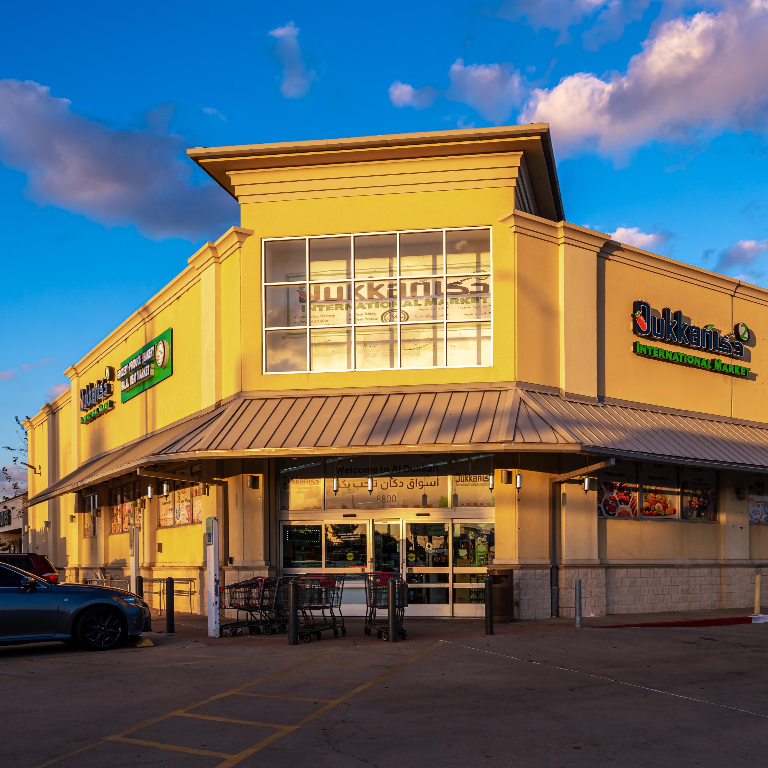 Interior of a supermarket with customers shopping near shelves stocked with packaged food, drinks, fresh produce, and a meat market counter in the background.