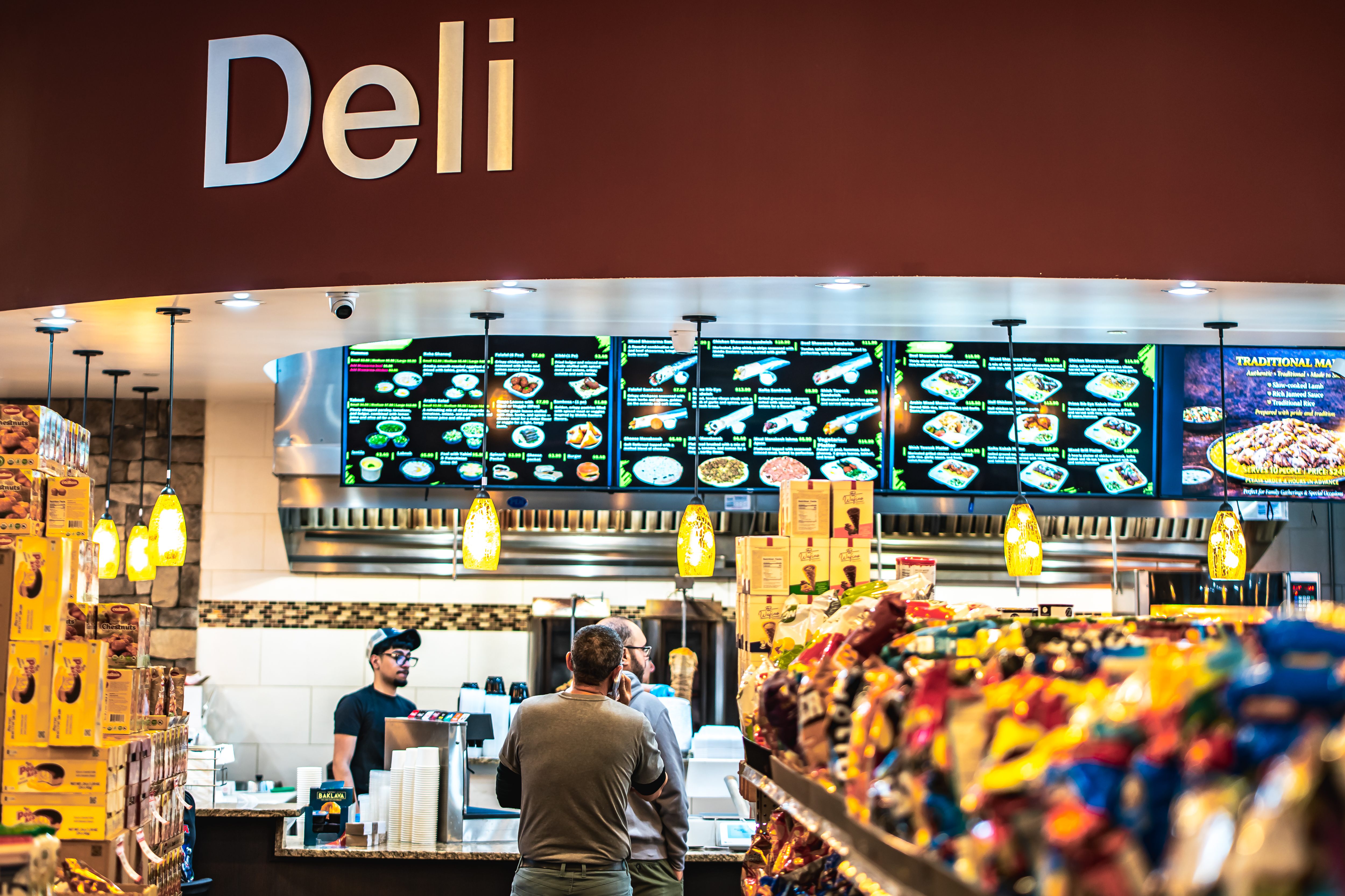Interior of a deli with a customer and employee at the counter, illuminated menu boards overhead, and shelves stacked with snacks.