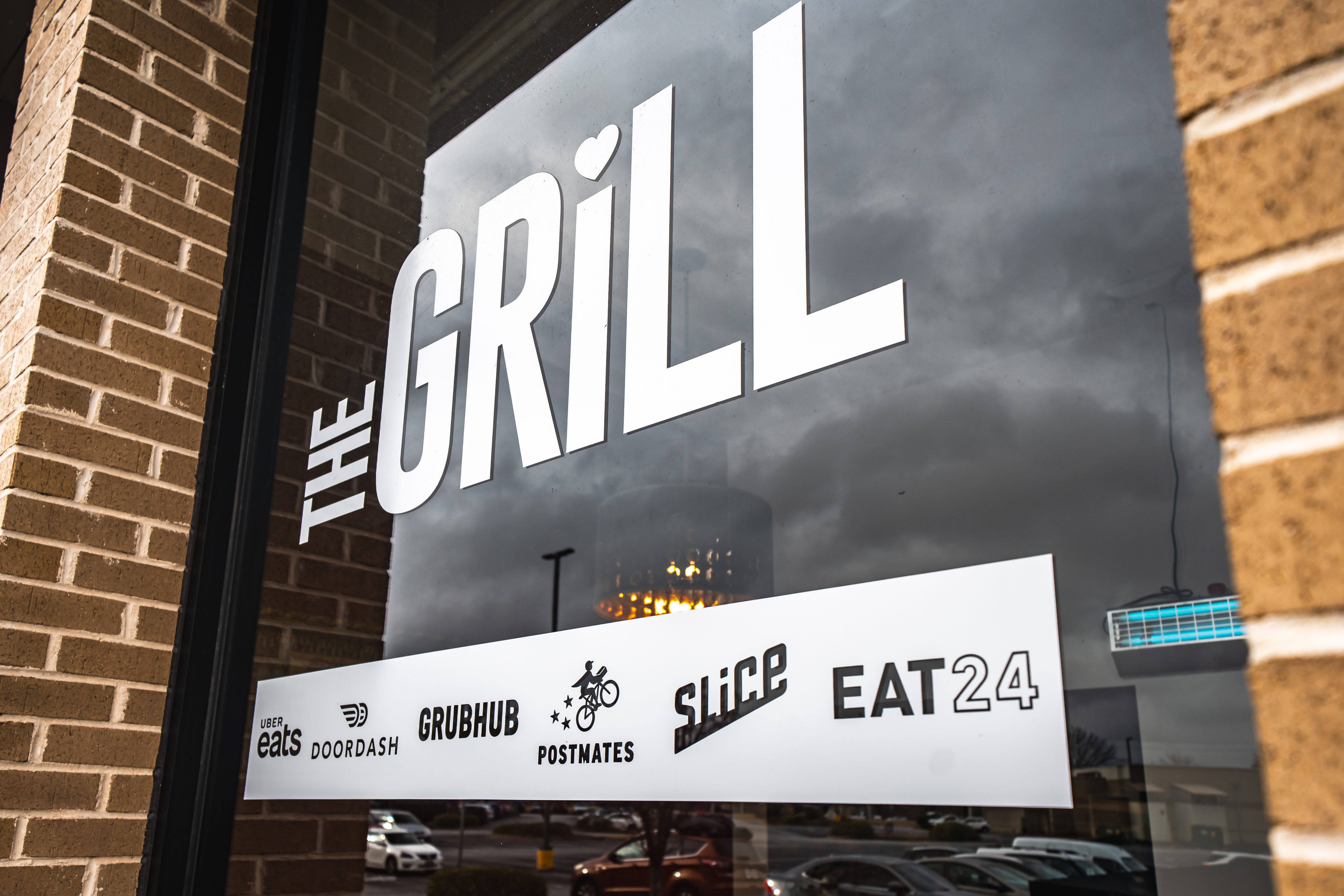Window sign for The Grill restaurant showing delivery service logos including Uber Eats, DoorDash, Grubhub, Postmates, Slice, and Eat24.