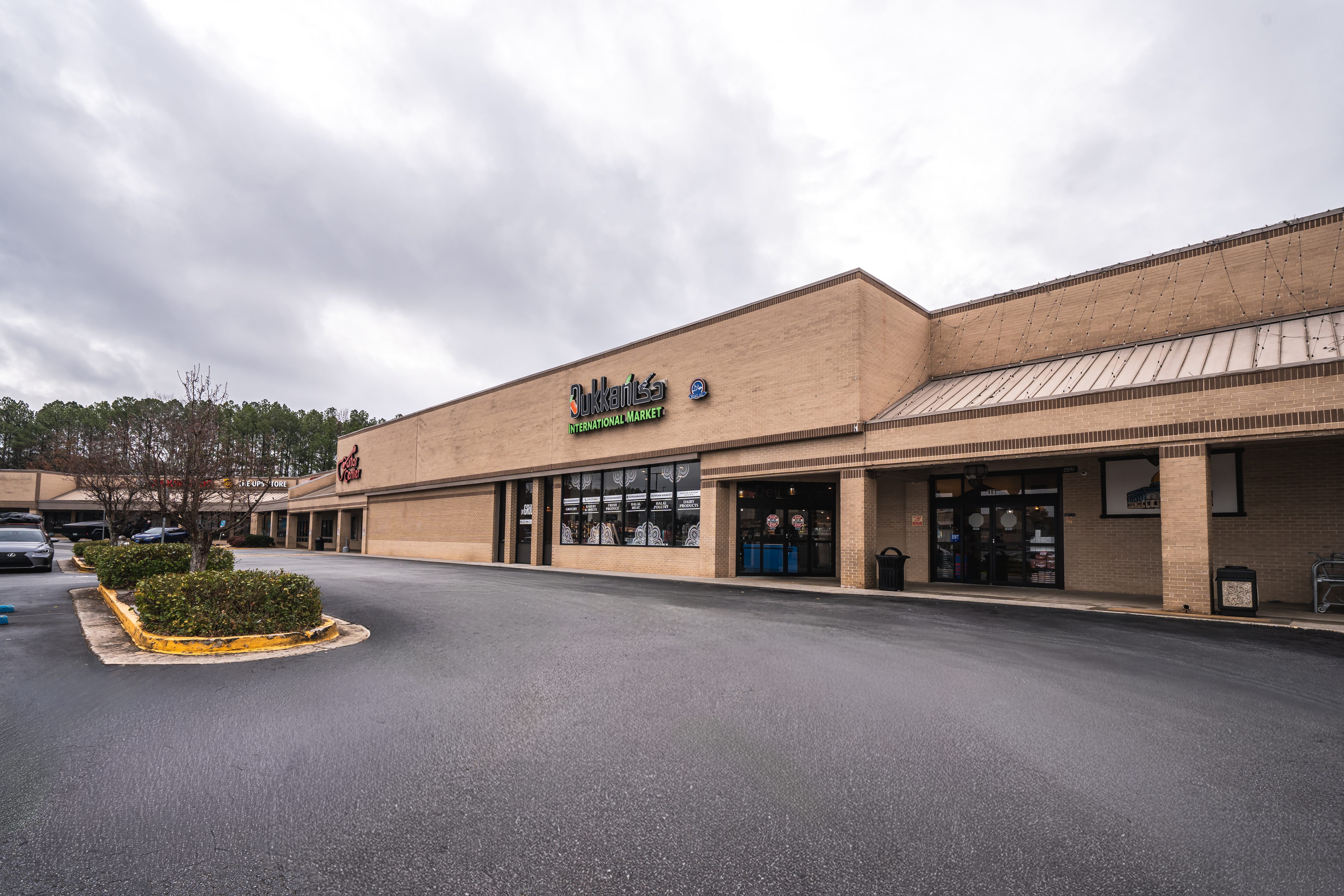 Exterior view of a beige brick strip mall with storefronts including Sukkan Lao International Market under a cloudy sky.