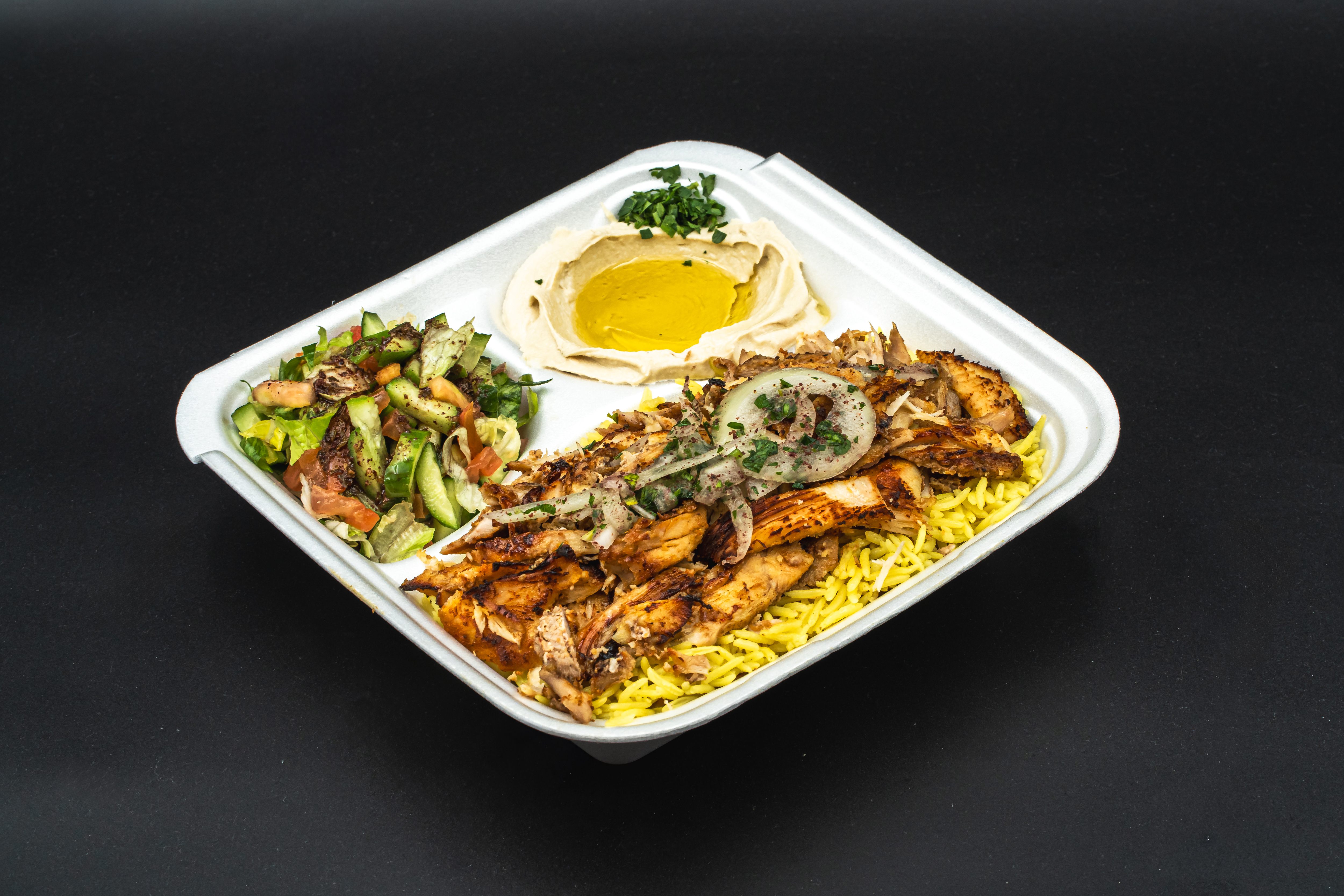 Takeout container with grilled chicken over yellow rice, hummus with olive oil and parsley, and fresh salad with cucumber and tomato.