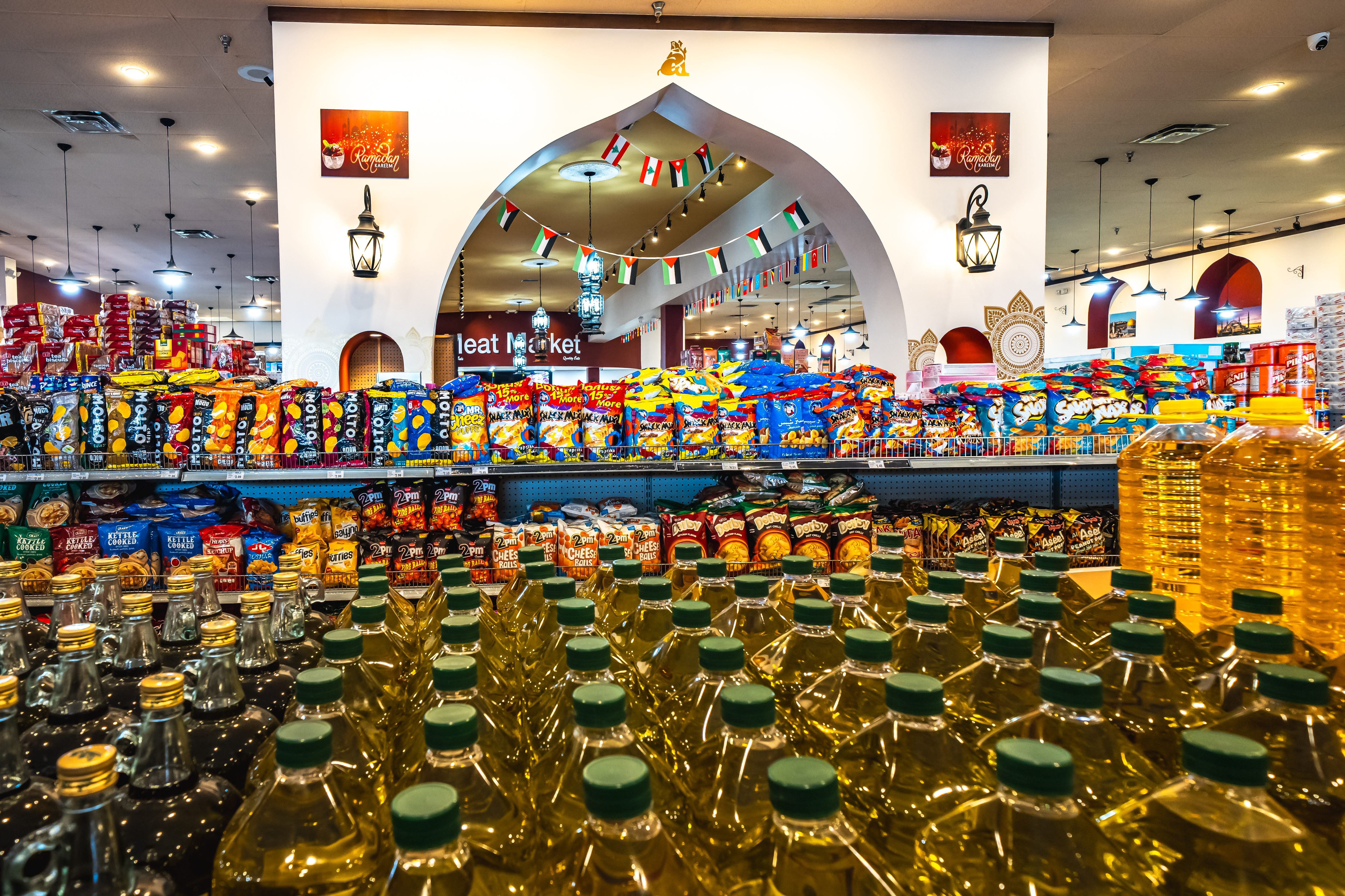 Supermarket aisle stocked with rows of cooking oil bottles in the foreground and various snack bags on shelves beneath an archway decorated with small hanging flags.