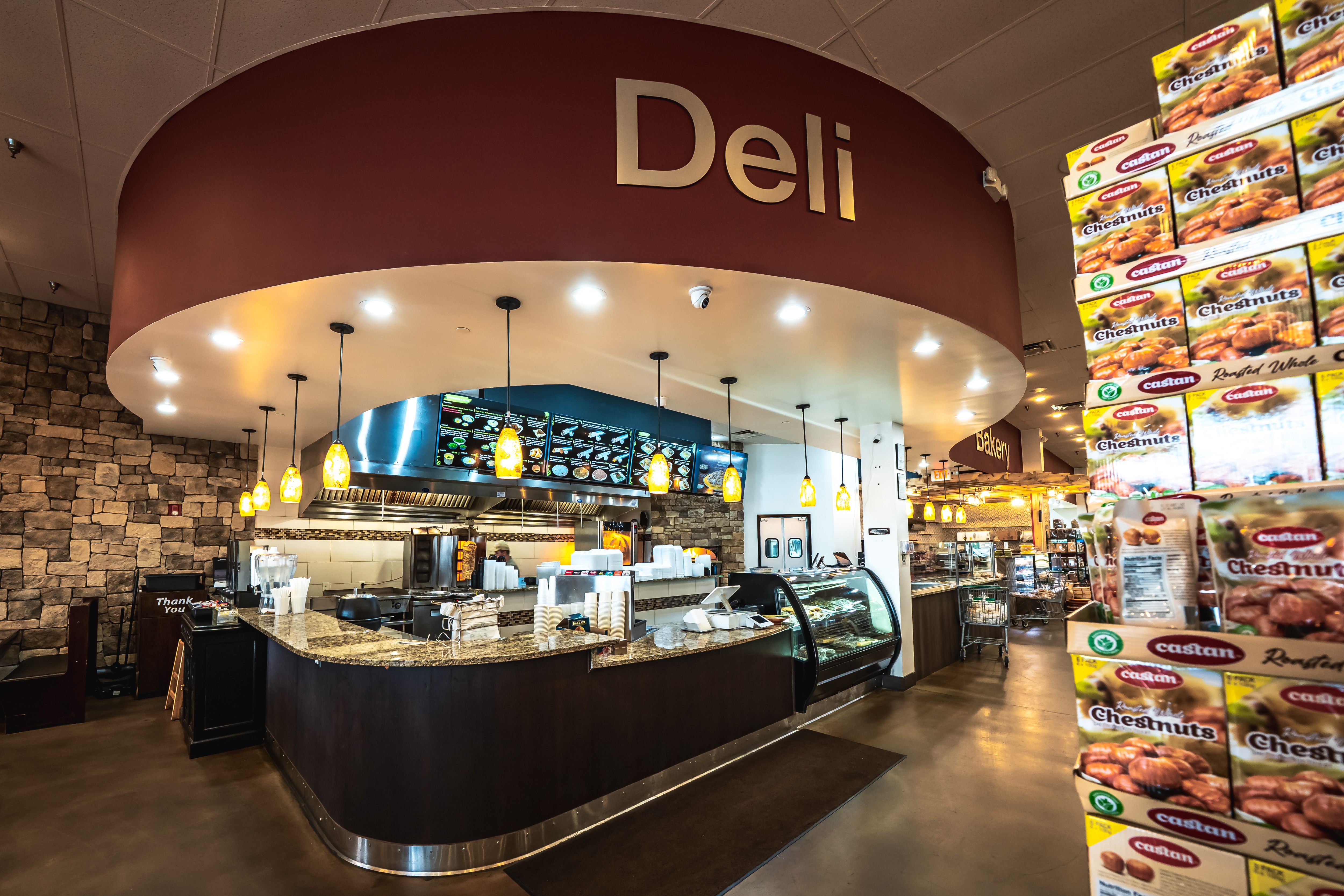 Interior of a deli counter with hanging pendant lights, digital menu screens, and a display case, next to shelves stocked with roasted chestnuts.