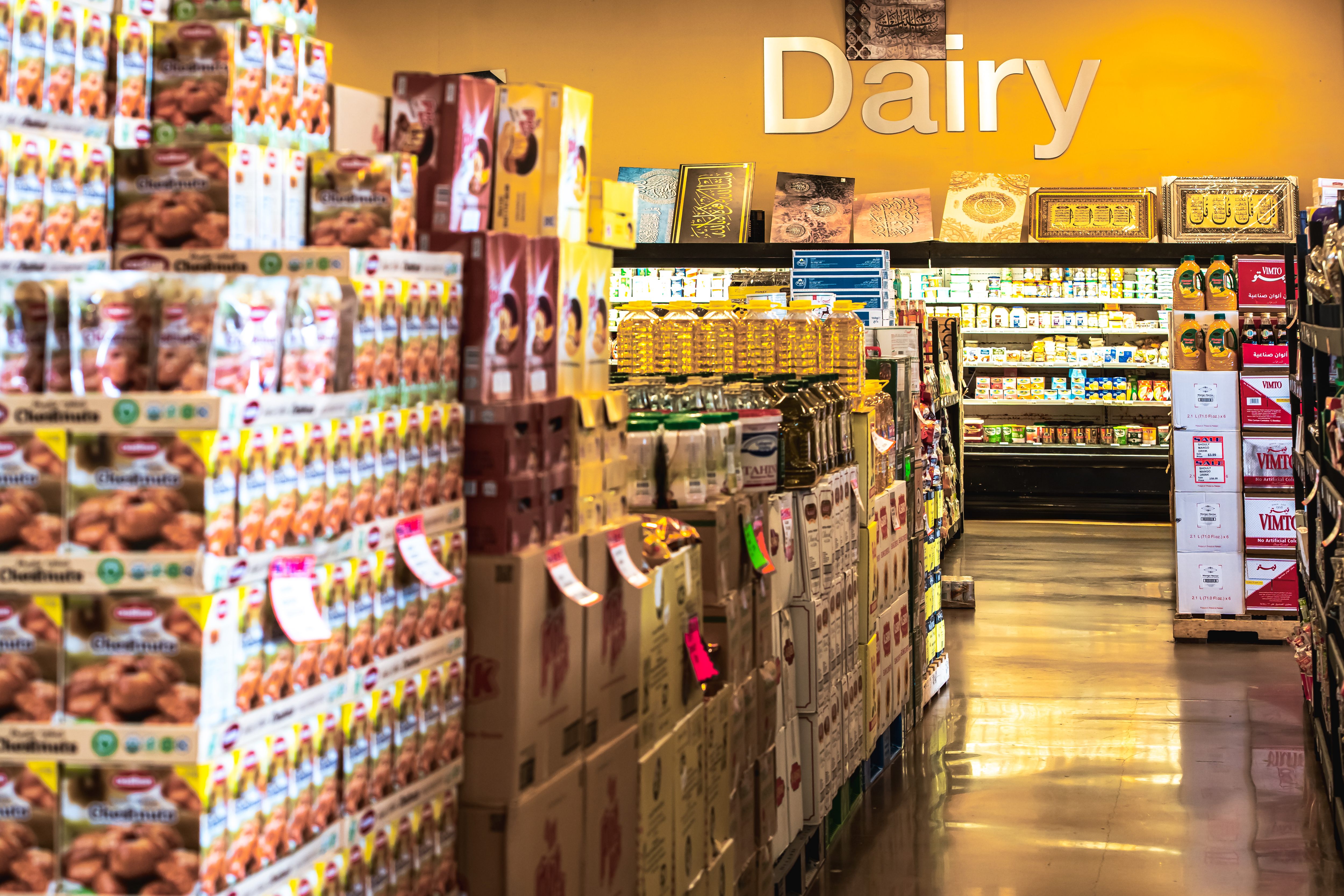 A grocery store aisle with stacks of packaged goods and a yellow wall sign labeled 'Dairy' above refrigerated dairy products.
