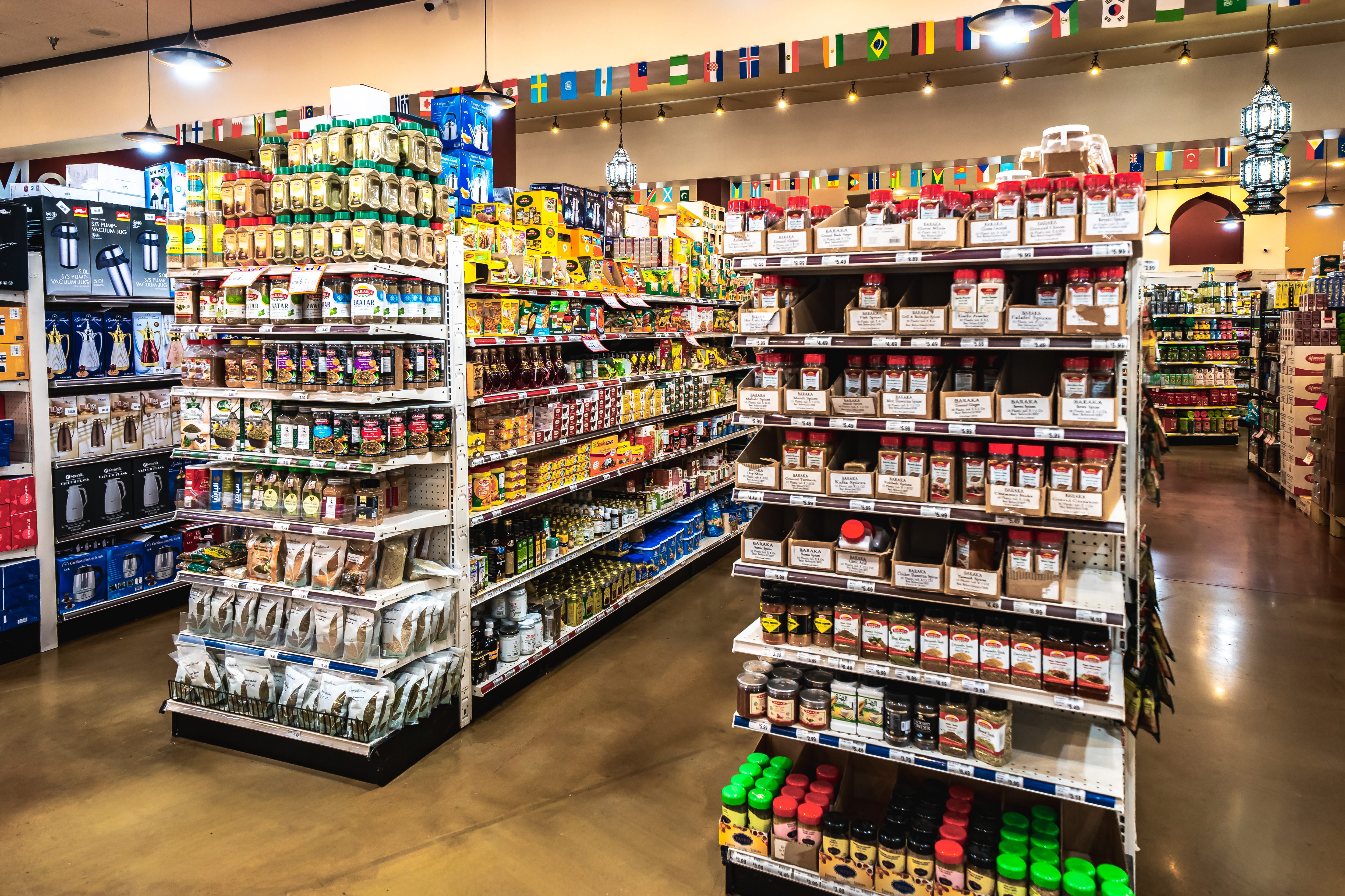 Interior of a Middle Eastern grocery store with a meat market at the back, shelves stocked with various packaged goods, fresh fruits, vegetables, and beverages, and customers shopping.
