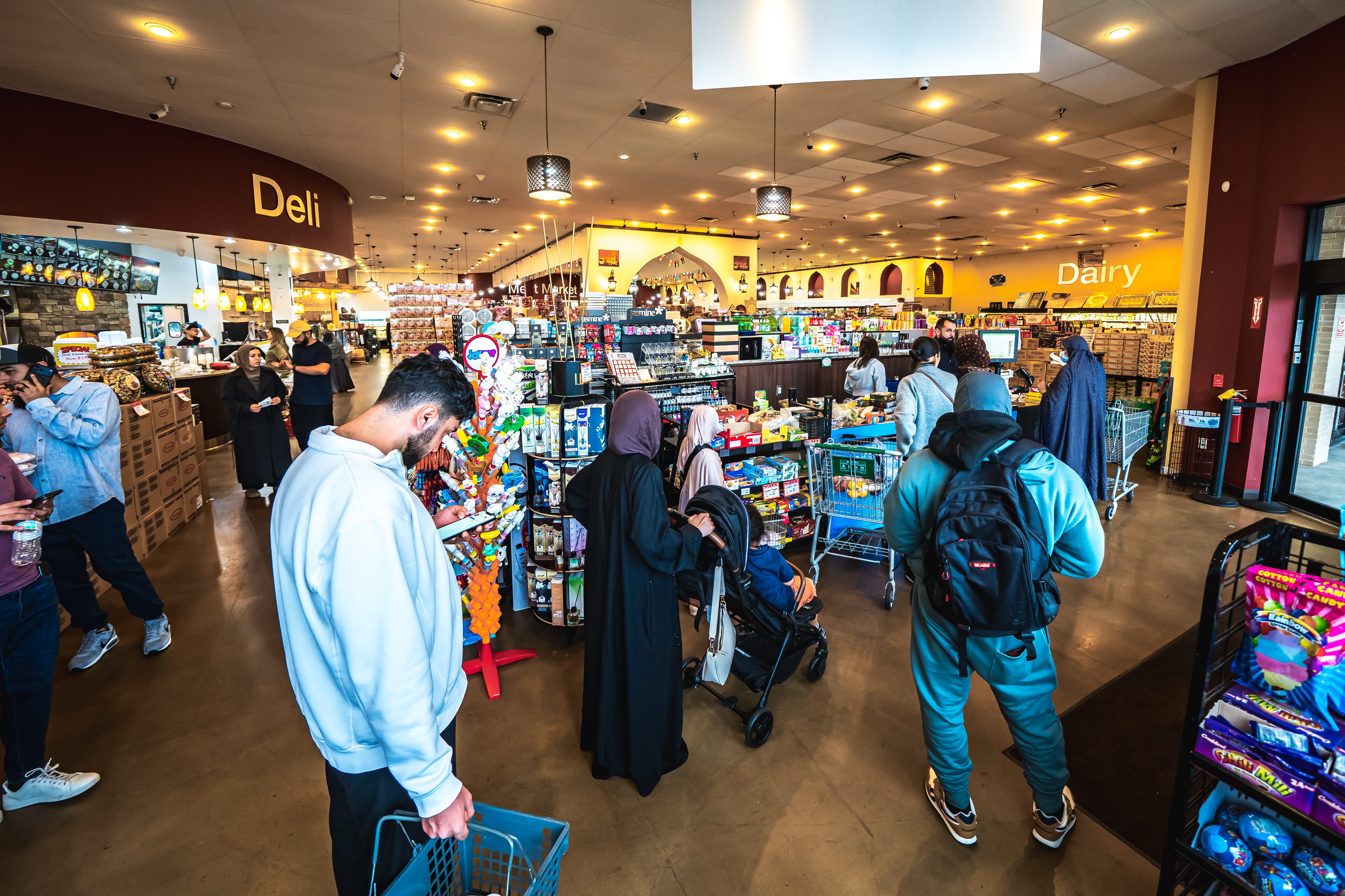 Shoppers standing in line with carts and baskets inside a brightly lit supermarket by deli and dairy sections.